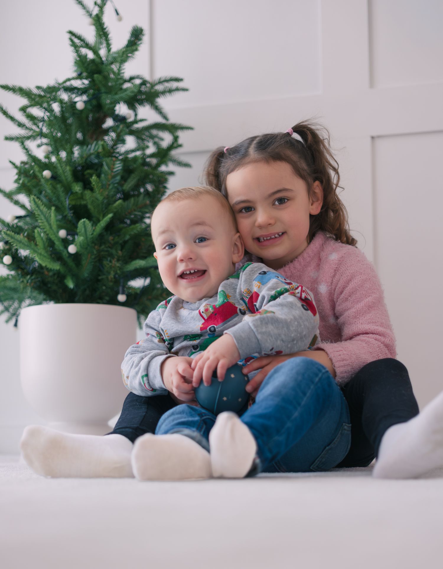 A smiling baby and older sister pose together near a small Christmas tree in a bright room.