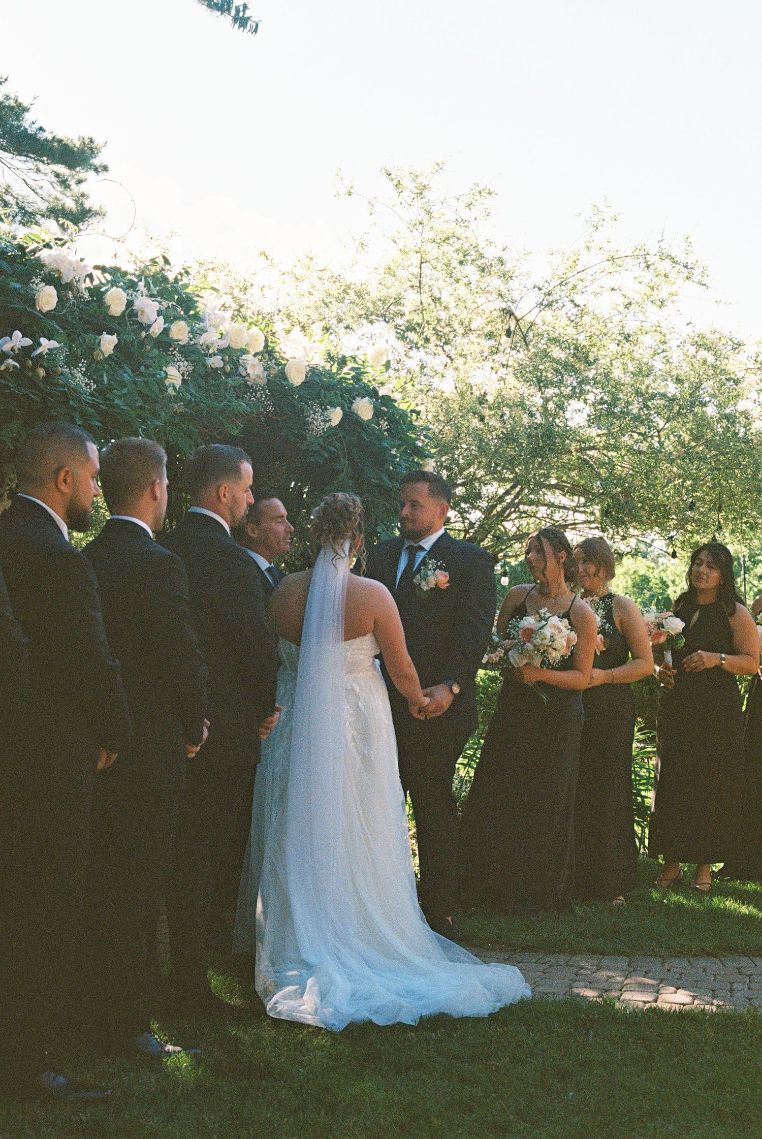 Outdoor wedding ceremony with bride and groom at floral arch, groomsmen in black suits, bridesmaids in black dresses.