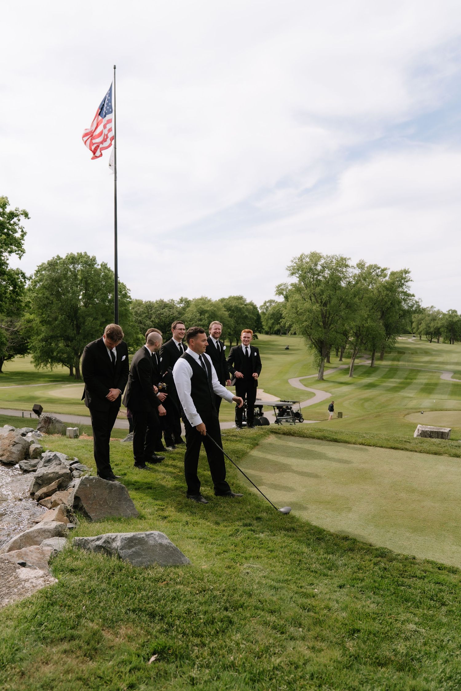 Groomsmen in black suits pose on a golf course hill near an American flag on a cloudy day.