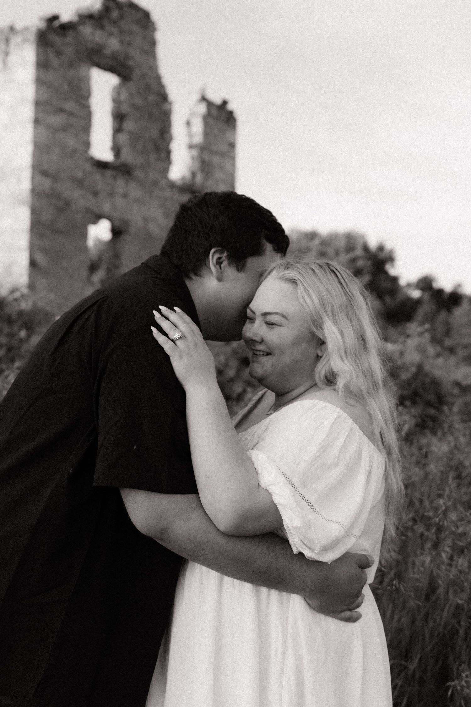 Black and white photo of couple embracing and laughing near rustic stone ruins outdoors.