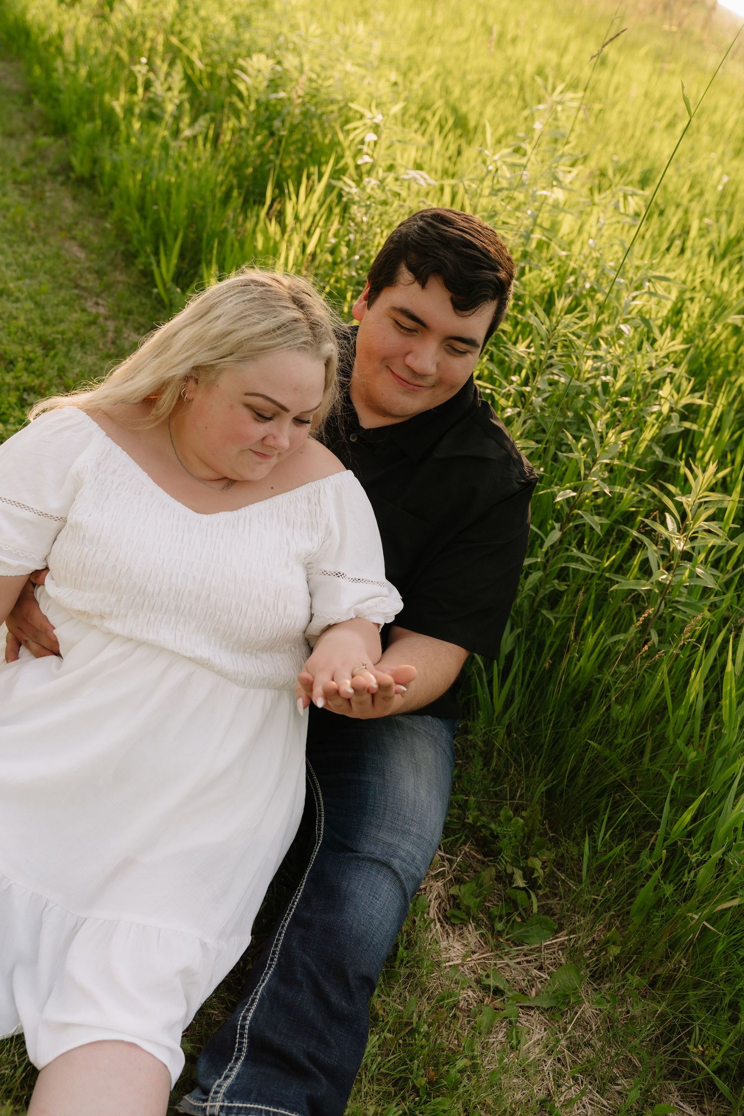 Couple sits in tall green grass, man in black shirt holding womans hand in white dress.
