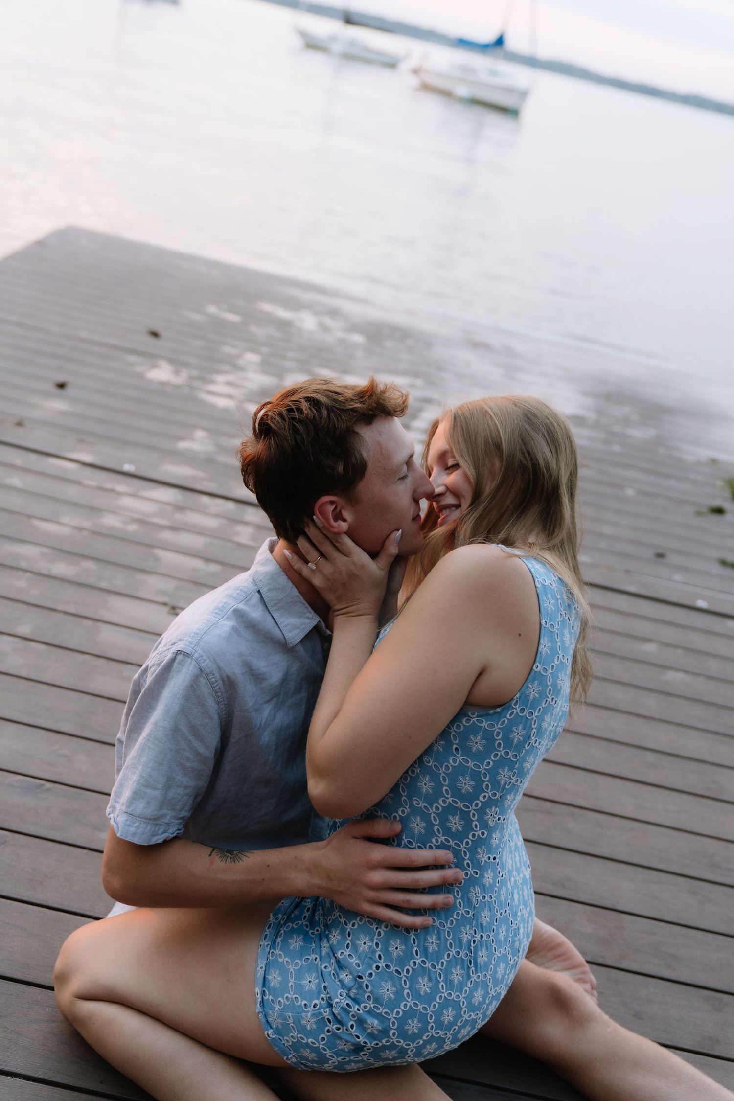 Couple sharing a romantic kiss on a wooden dock by the water, woman in blue dress embracing man in gray shirt.
