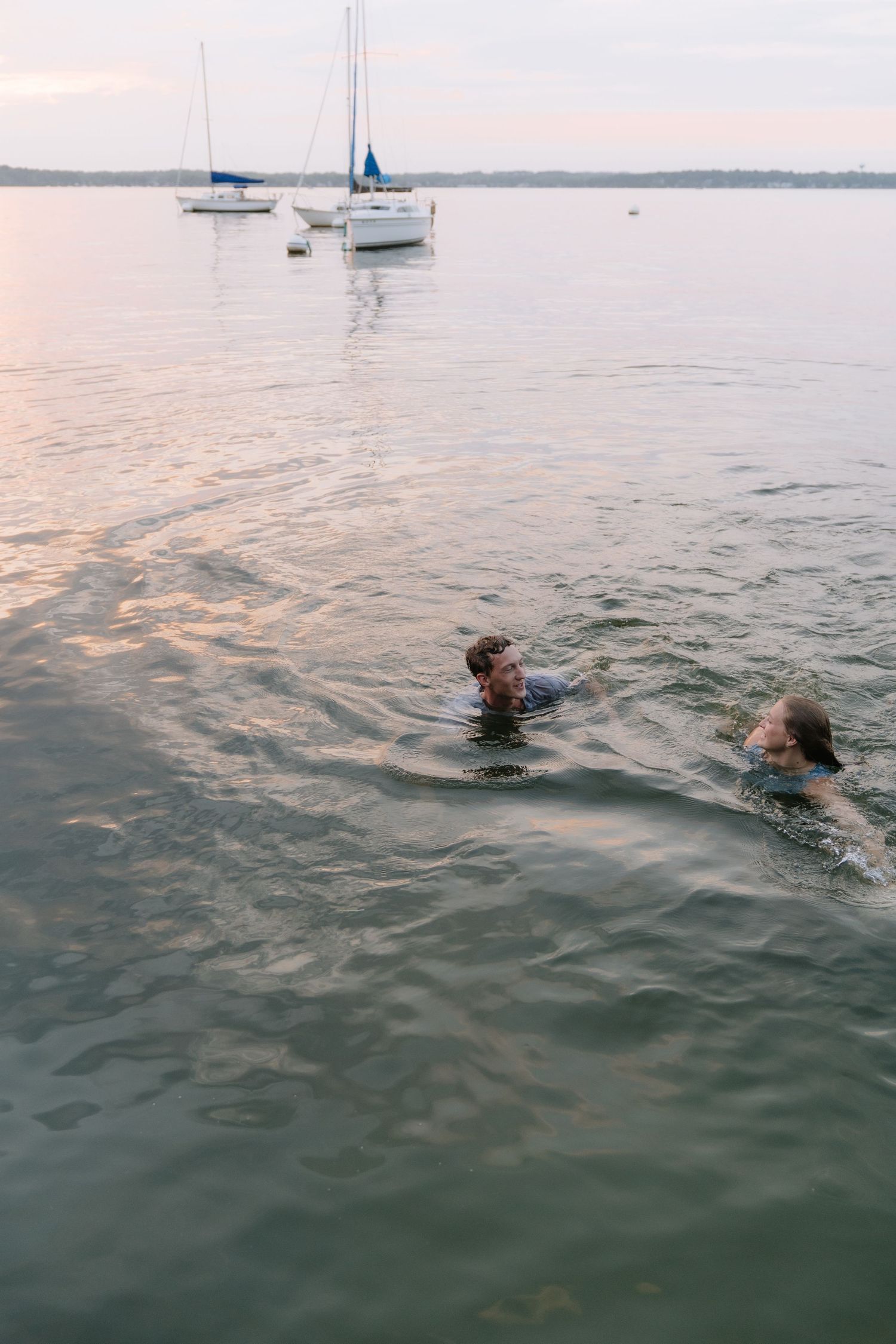 Two people swimming in calm lake waters at dusk with sailboats anchored in the background.