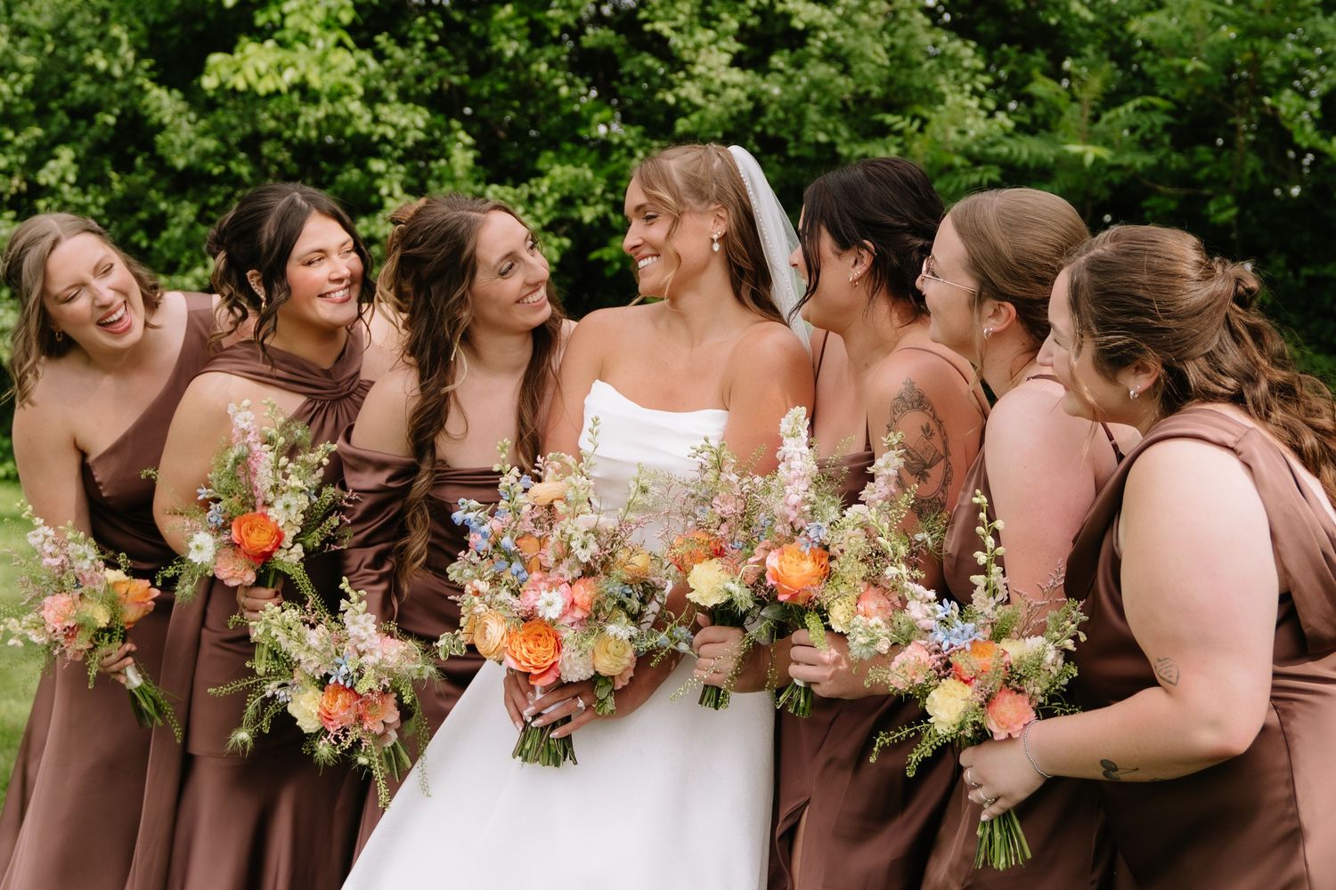 Bride in white gown laughing with bridesmaids in brown dresses holding colorful bouquets outdoors.
