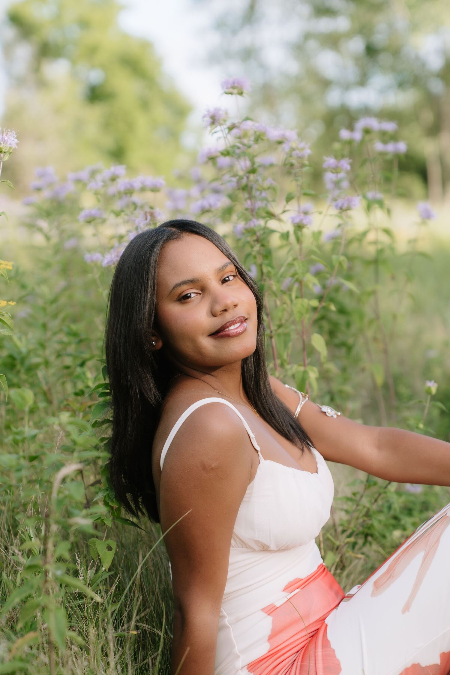 Young woman in white floral dress smiling among purple wildflowers in a lush green outdoor setting.