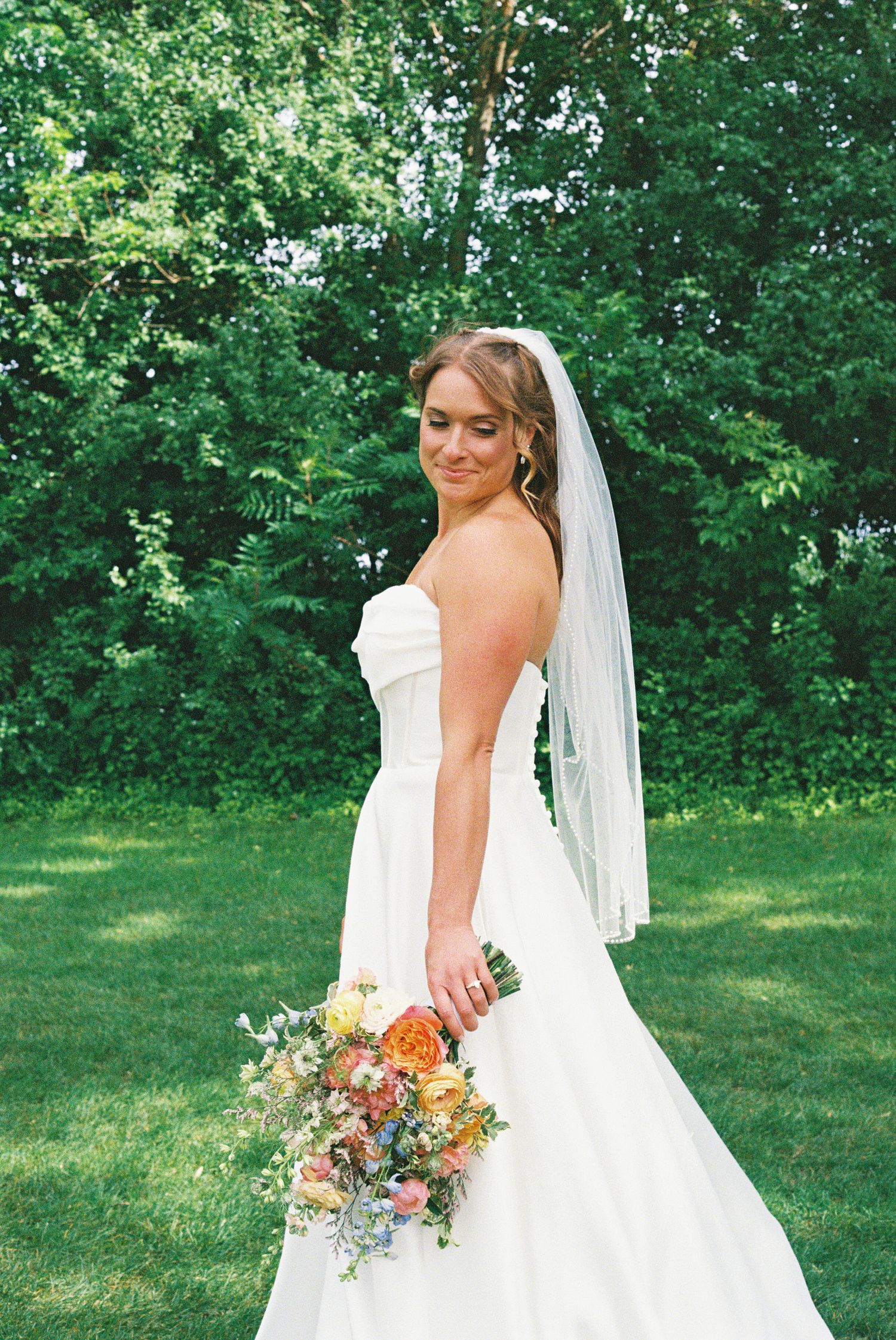 Bride in strapless white gown and veil holds colorful bouquet, smiling over shoulder on green lawn.
