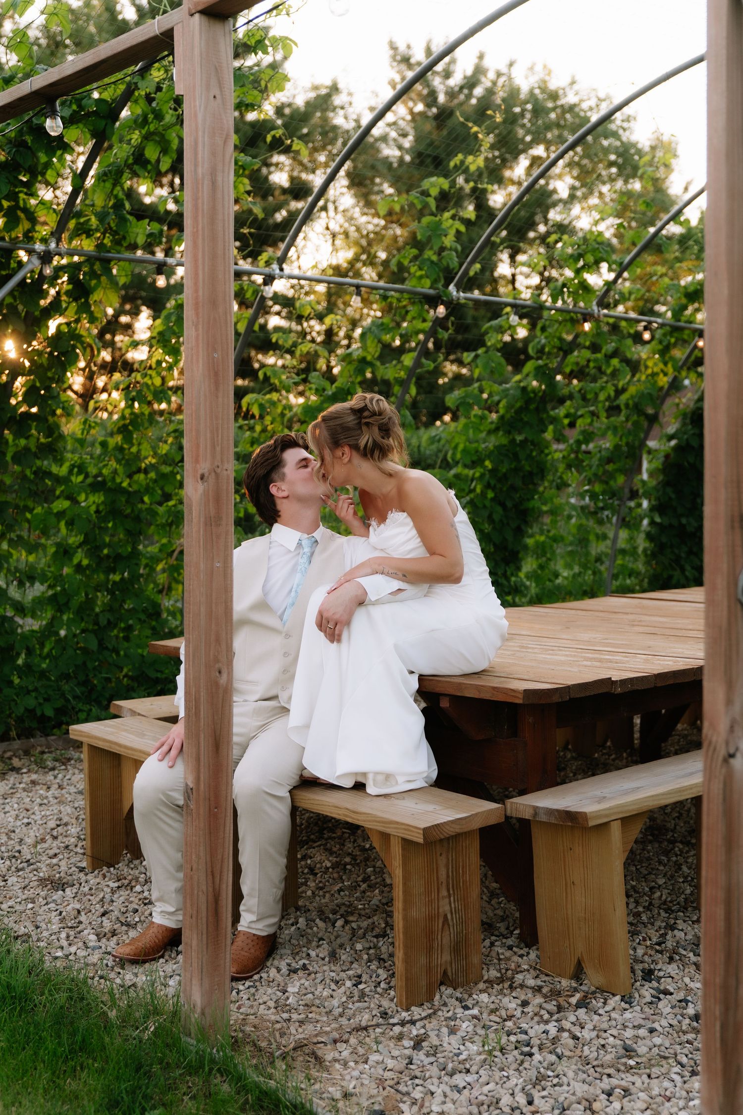 Bride leans in to kiss groom seated on wooden bench under pergola with string lights at dusk.