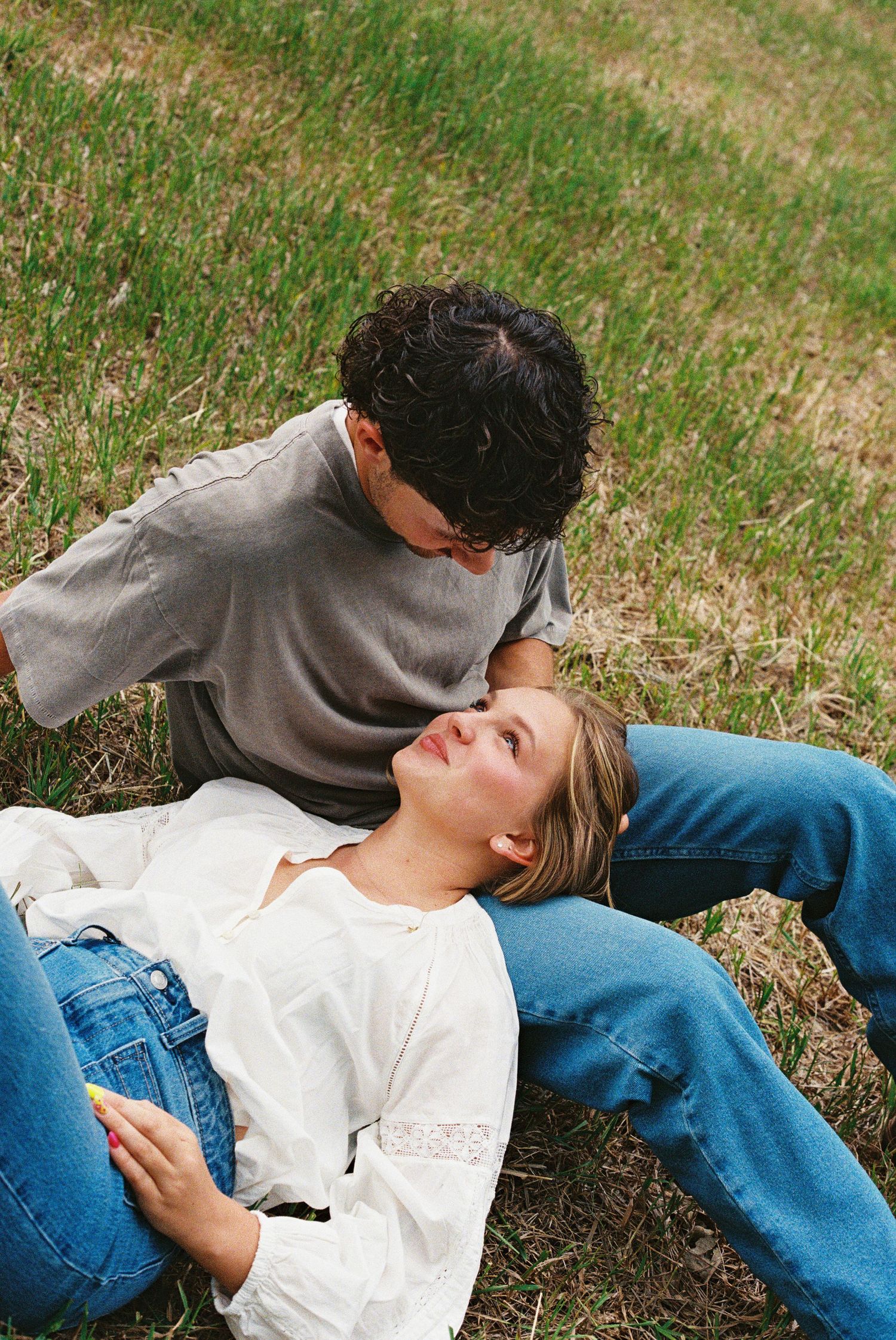 A young couple relaxing on green grass, woman lying down in white blouse and jeans, man sitting beside her in gray tee.