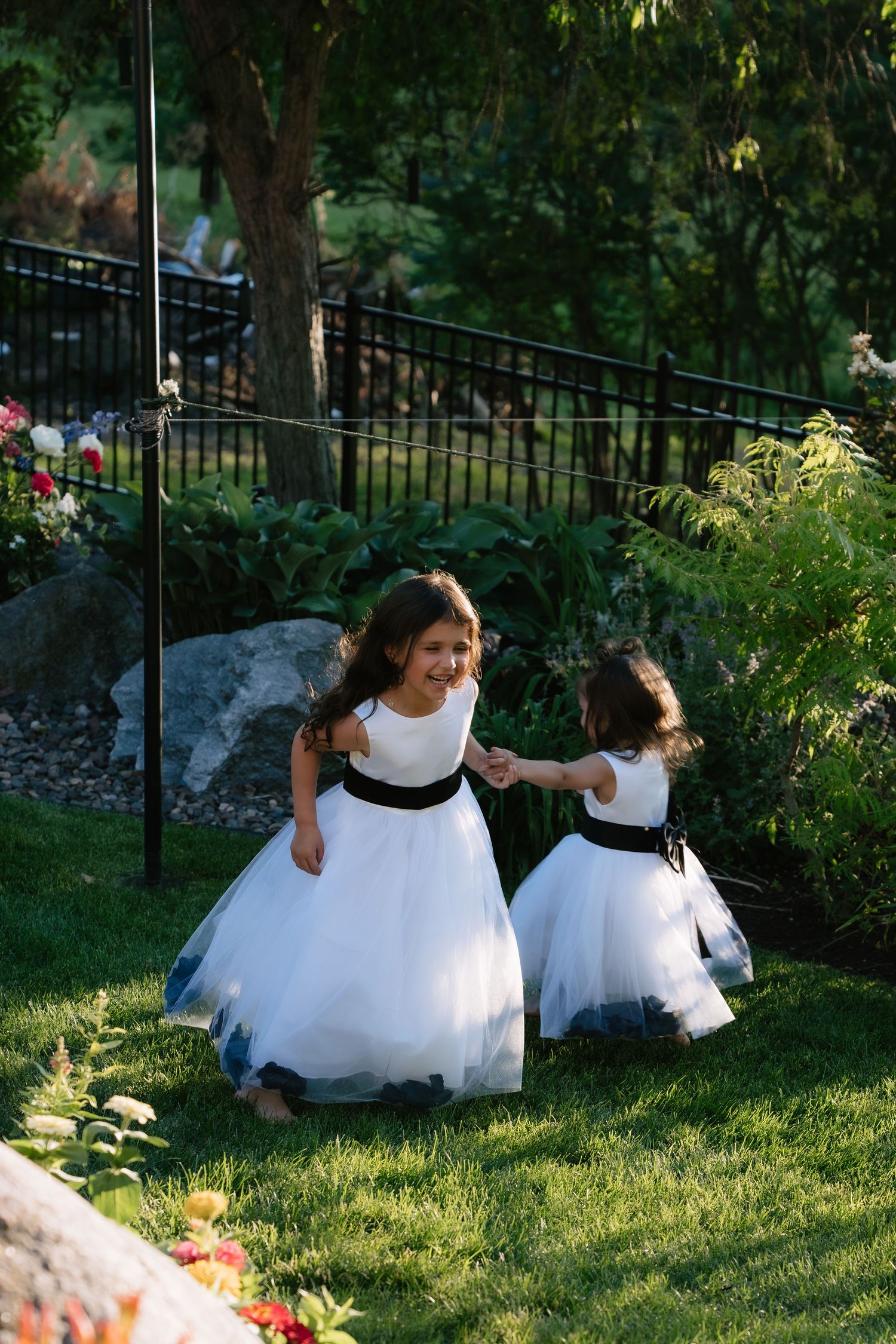 Two flower girls in white and black dresses playing together on green grass at an outdoor wedding venue.