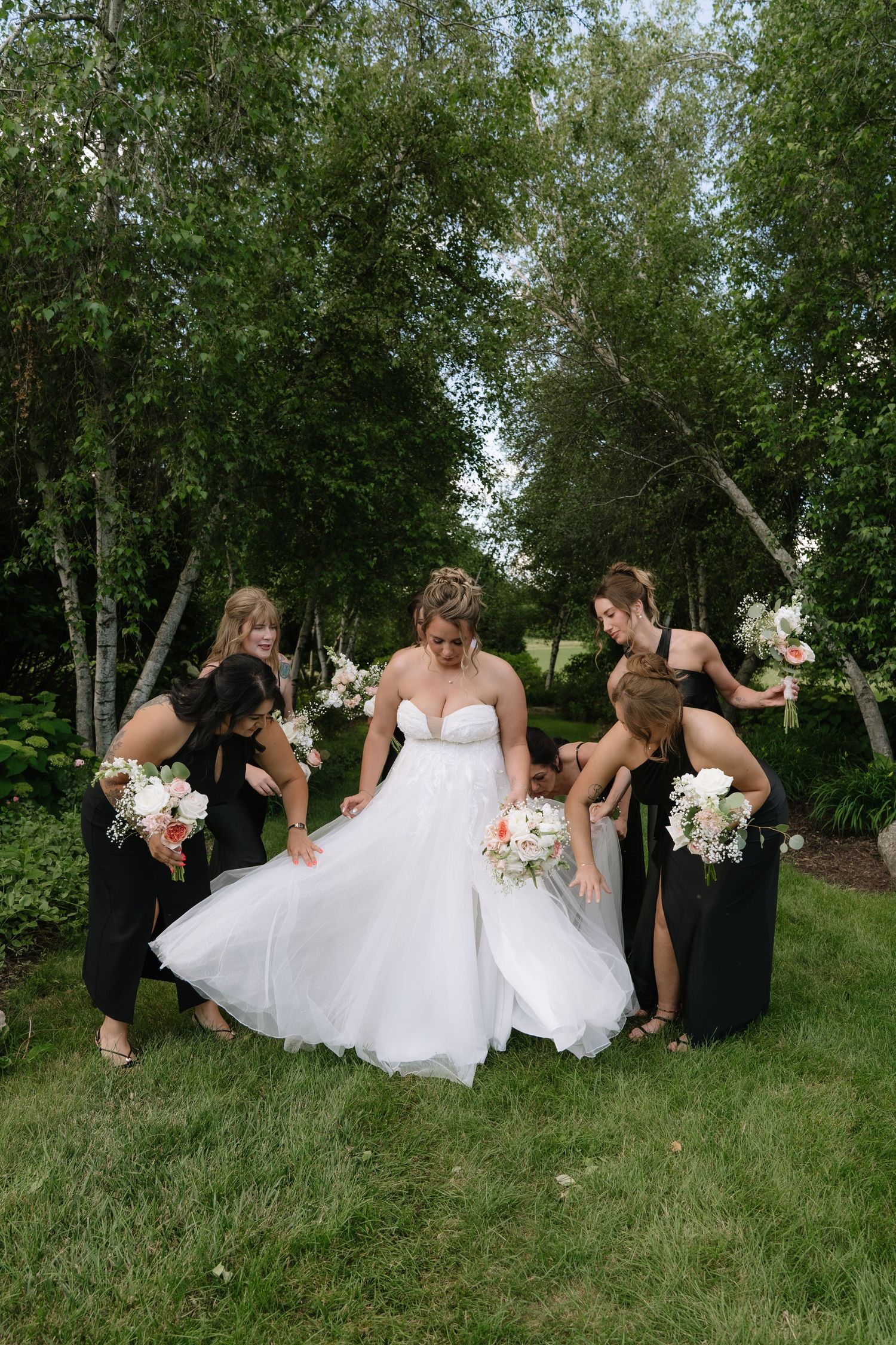 Bride in white gown surrounded by bridesmaids in black dresses adjusting her skirt outdoors among lush green trees.