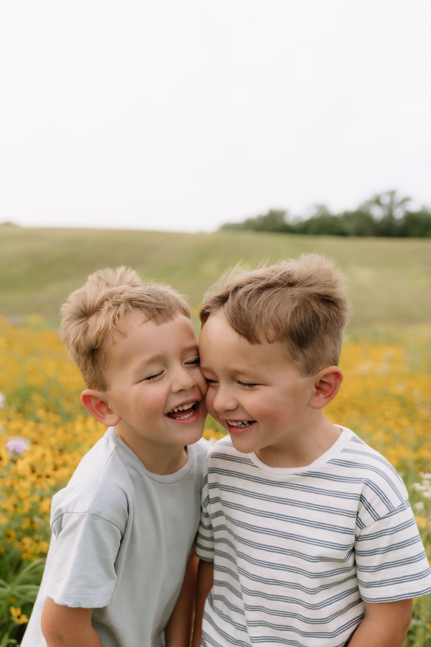 Two laughing blonde boys in t-shirts share a joyful moment together in a sunny wildflower field.