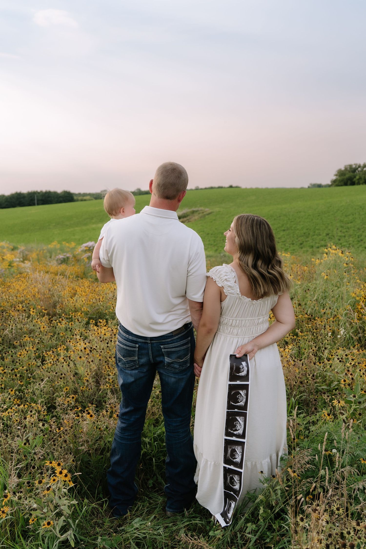 A couple stands in a flower field, man holding baby, woman in white dress displaying ultrasound photos at sunset.