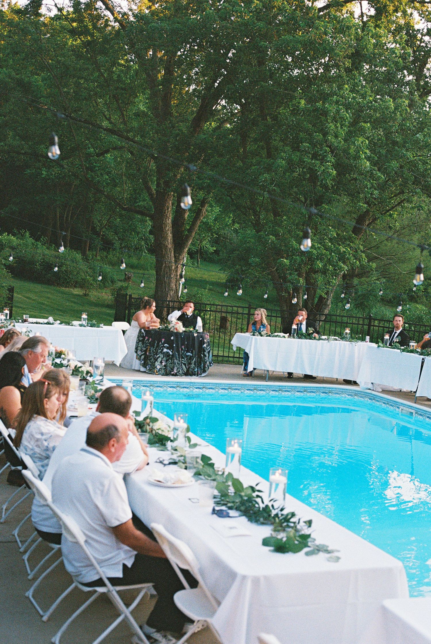 Elegant outdoor wedding reception dinner beside a blue swimming pool, with white tablecloths, candles, and string lights in trees.