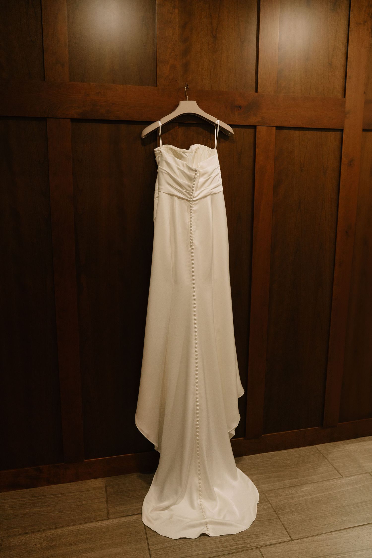 Elegant strapless white wedding gown with button back detail hanging on wooden wall.
