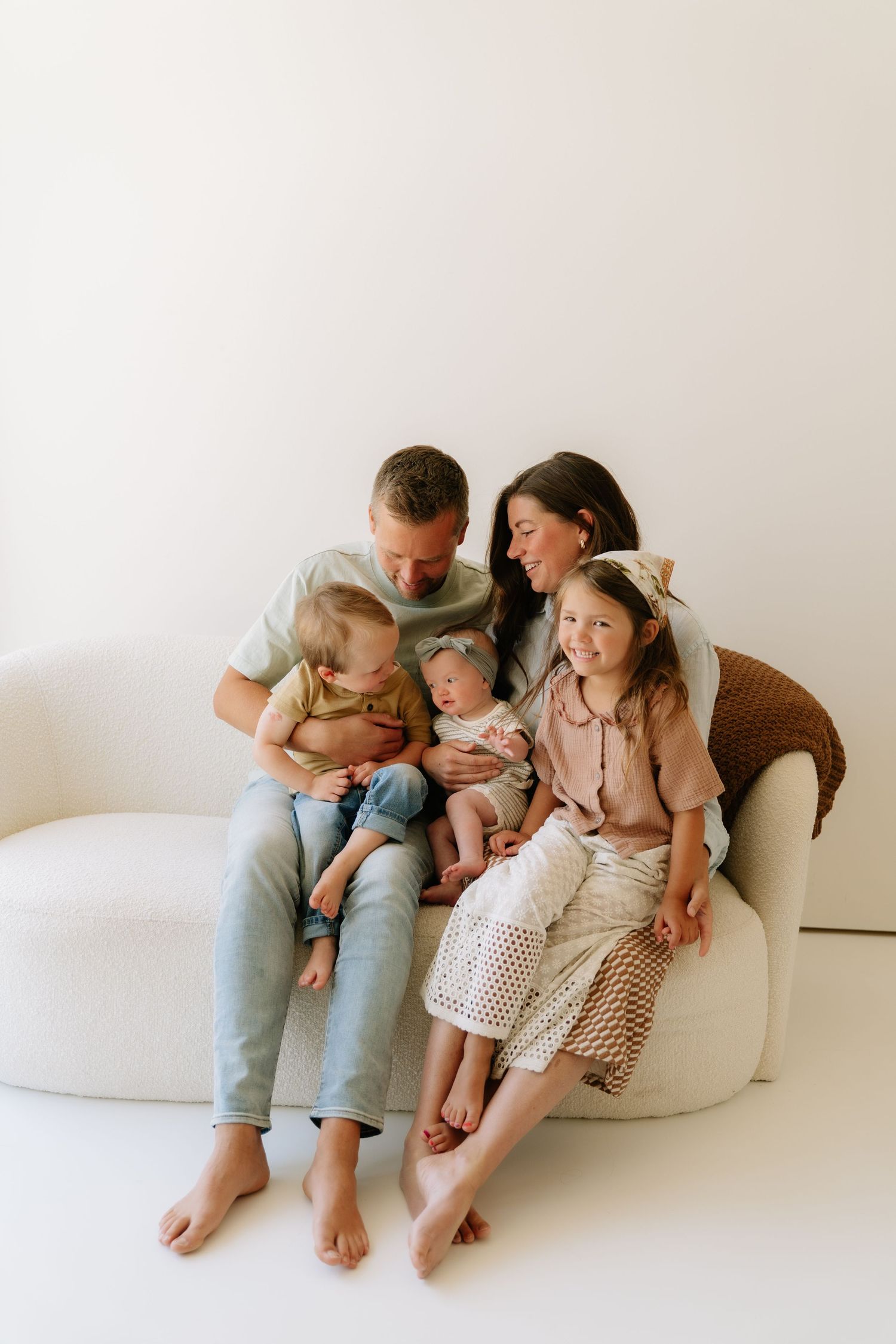 A happy family of five sits on a white sofa, parents smiling at their newborn baby with two young daughters beside them.