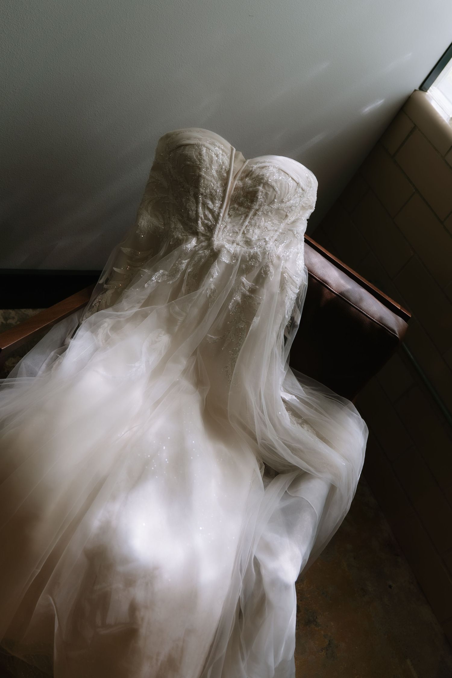 Elegant lace wedding dress draped over a wooden chair, featuring a strapless bodice and flowing tulle skirt in soft natural light.