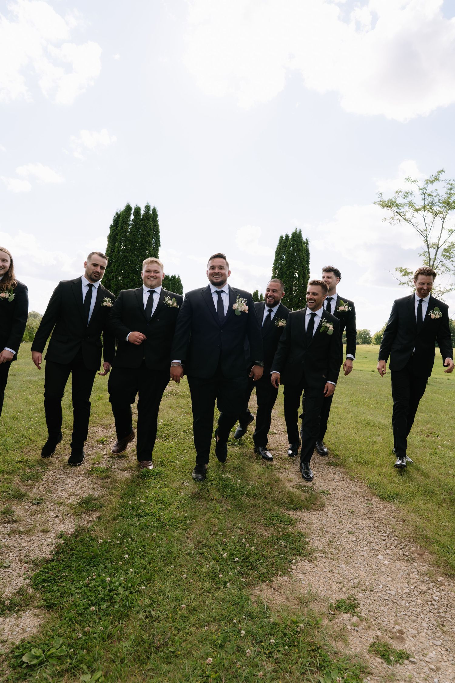 Groom and groomsmen in black suits walk confidently along a gravel path at an outdoor wedding venue.