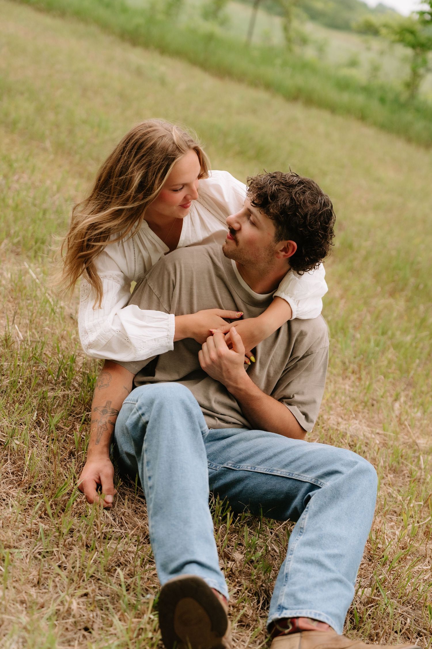A couple sits in a grassy field, gazing into each other's eyes and holding hands in a romantic outdoor moment.
