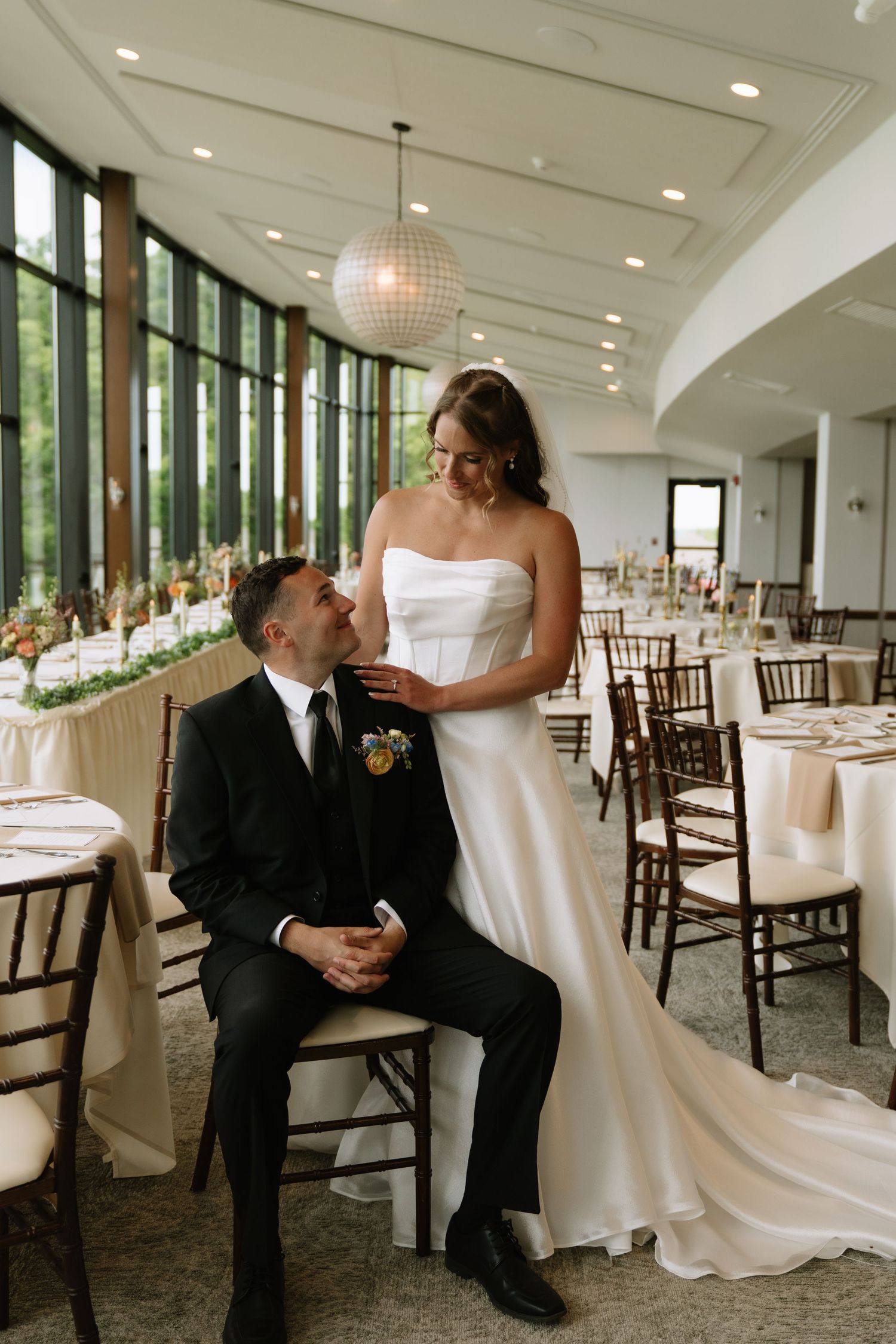 Bride in white strapless gown stands beside seated groom in black suit at elegant wedding reception venue.