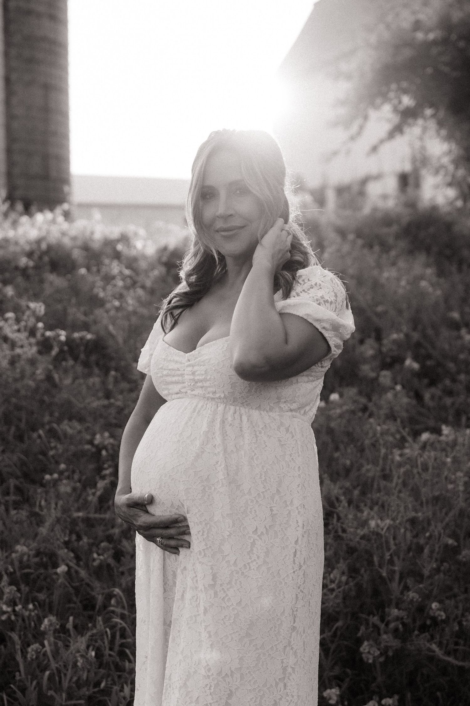 Pregnant woman in white lace dress poses in a sunlit field during a maternity photo shoot.