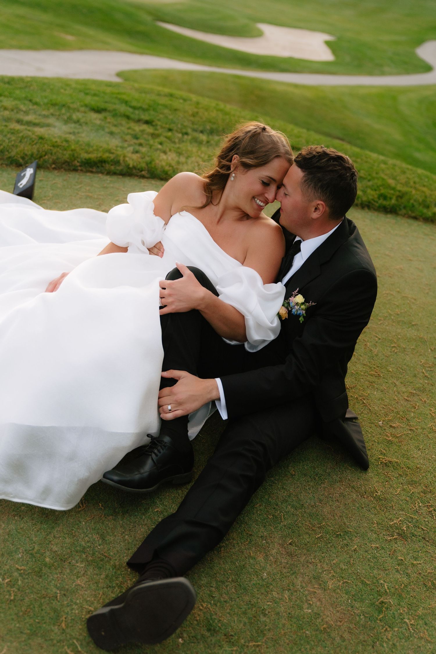 Bride and groom sitting on green golf course grass, sharing a romantic moment in wedding attire.