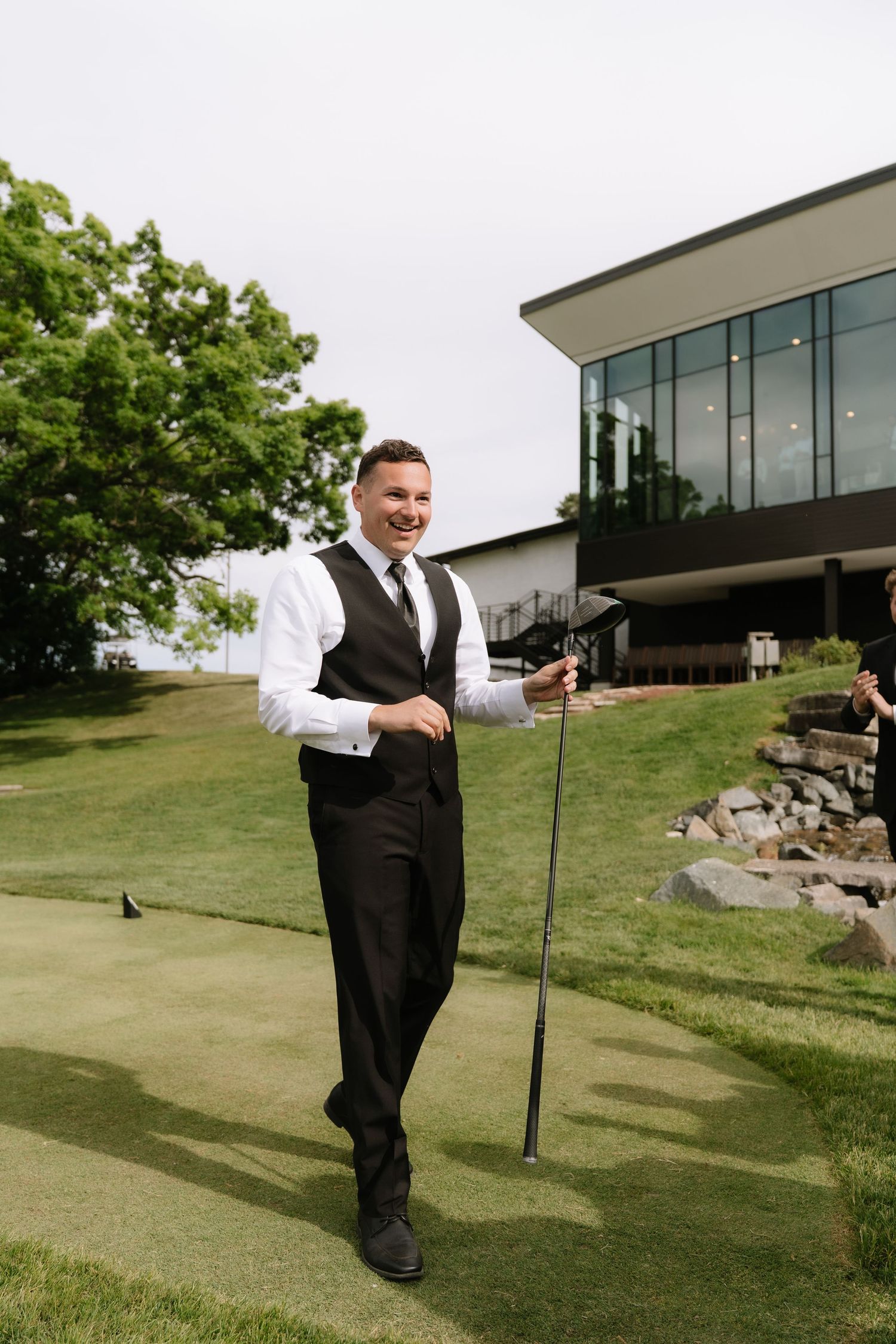 Smiling groom in black suit and vest holds golf club on green course near elegant venue building.