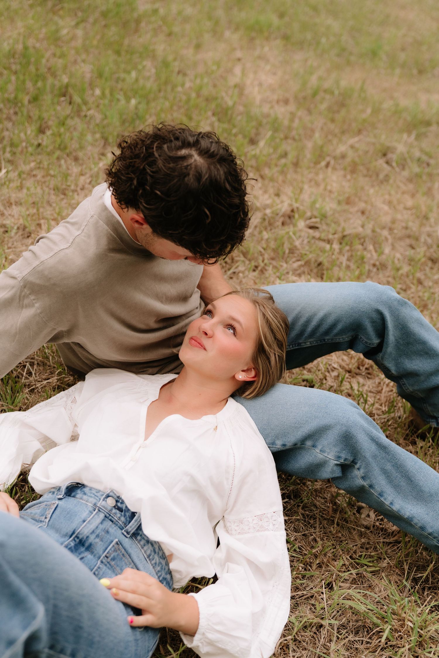 A couple relaxes on grass, woman in white blouse lying back as man leans in to kiss her forehead.