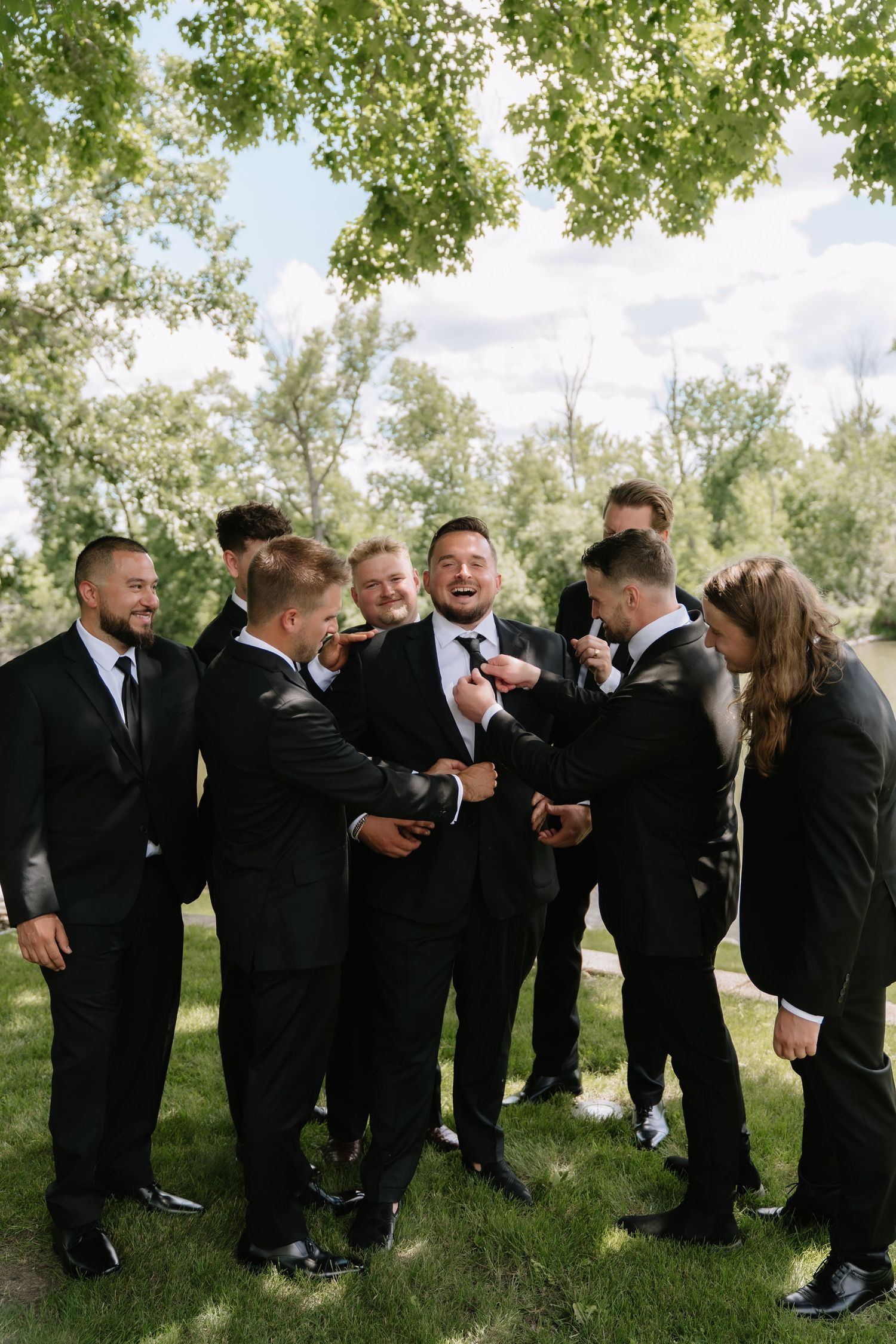 Groom surrounded by groomsmen in black suits laughing and adjusting ties outdoors under green trees on wedding day.