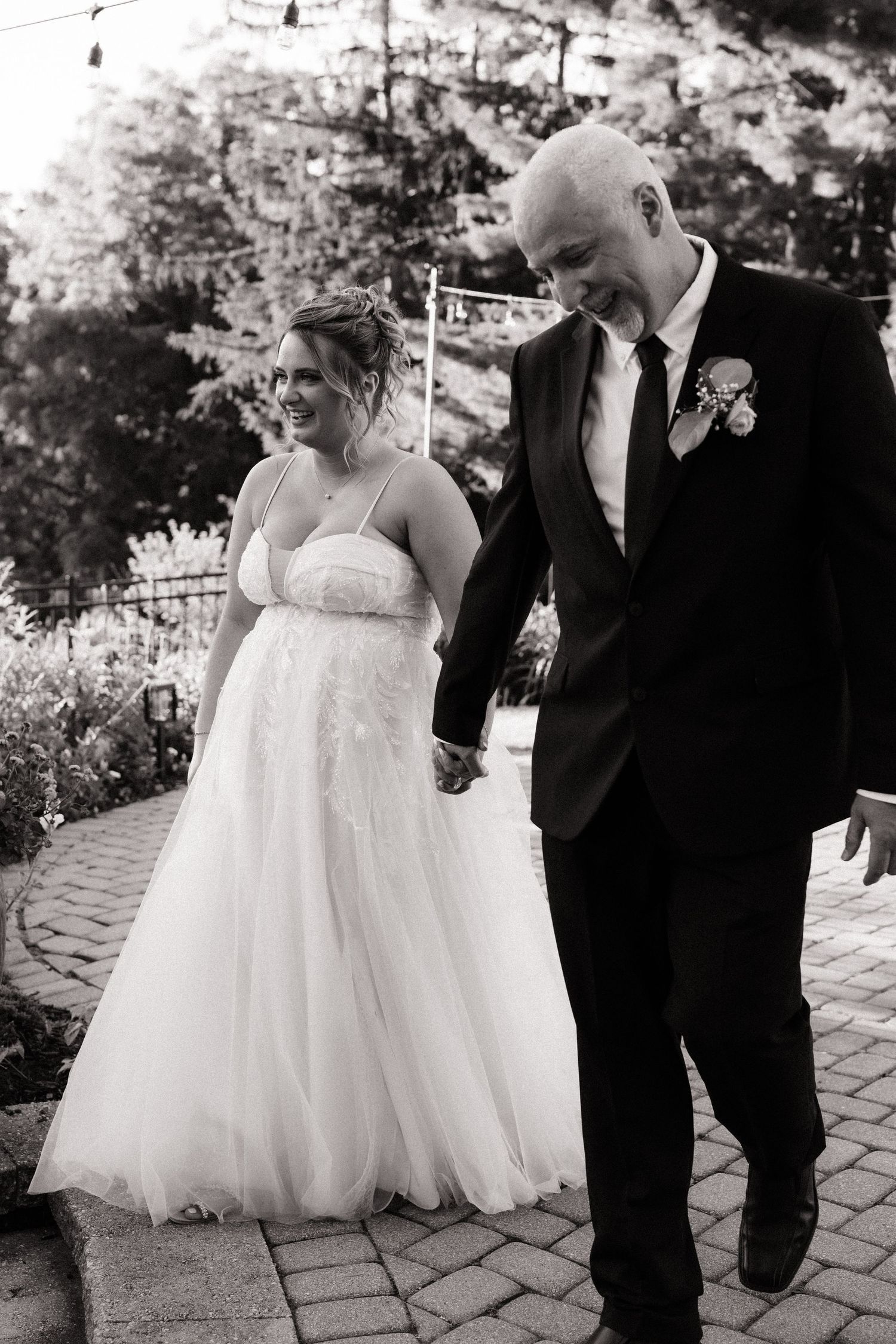 Black and white photo of a smiling bride in a white gown holding hands with a groom in a black suit at an outdoor wedding.