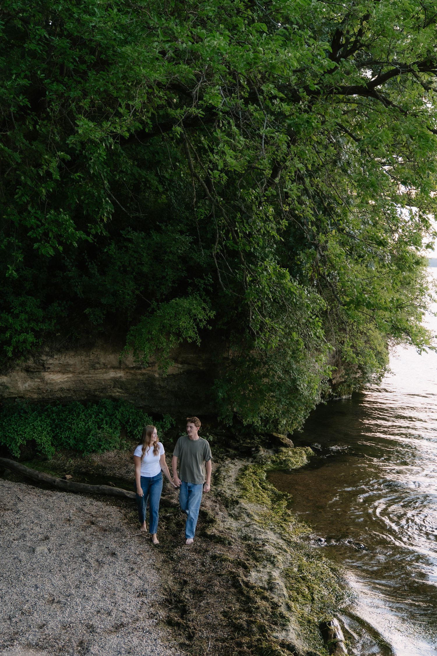 Couple holding hands walking along a rocky riverbank surrounded by lush green trees and limestone cliffs.