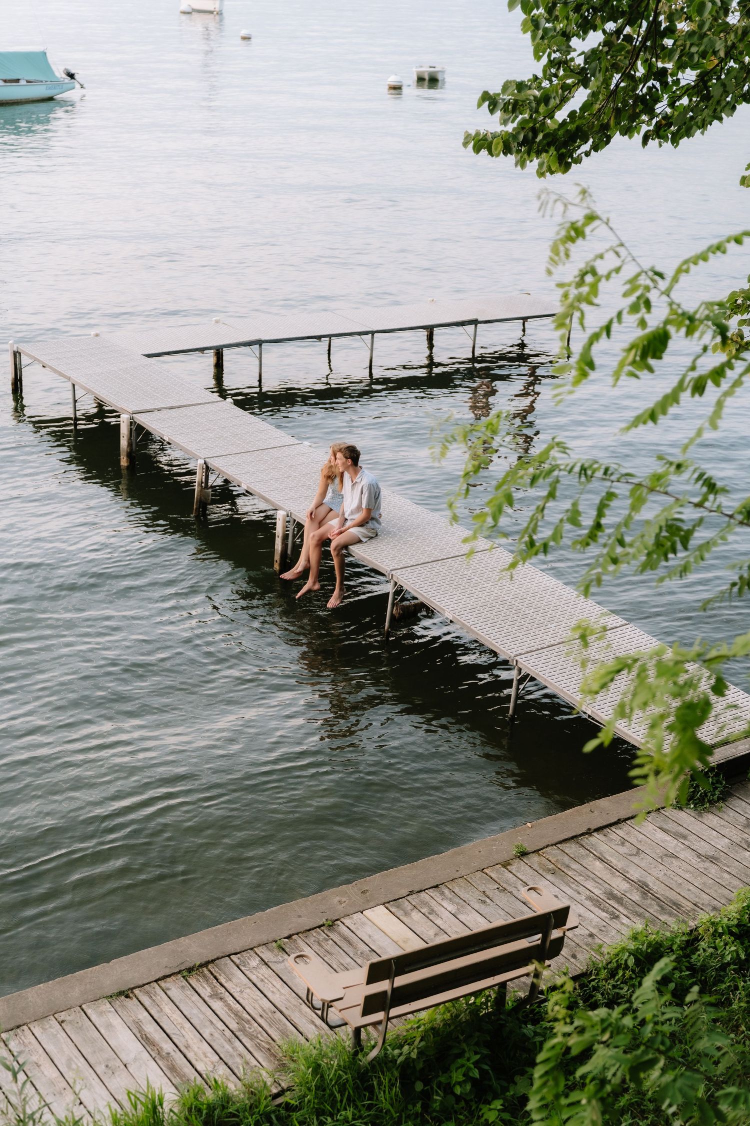 Couple sitting close together on a wooden dock over calm lake water, with a bench and green trees nearby.