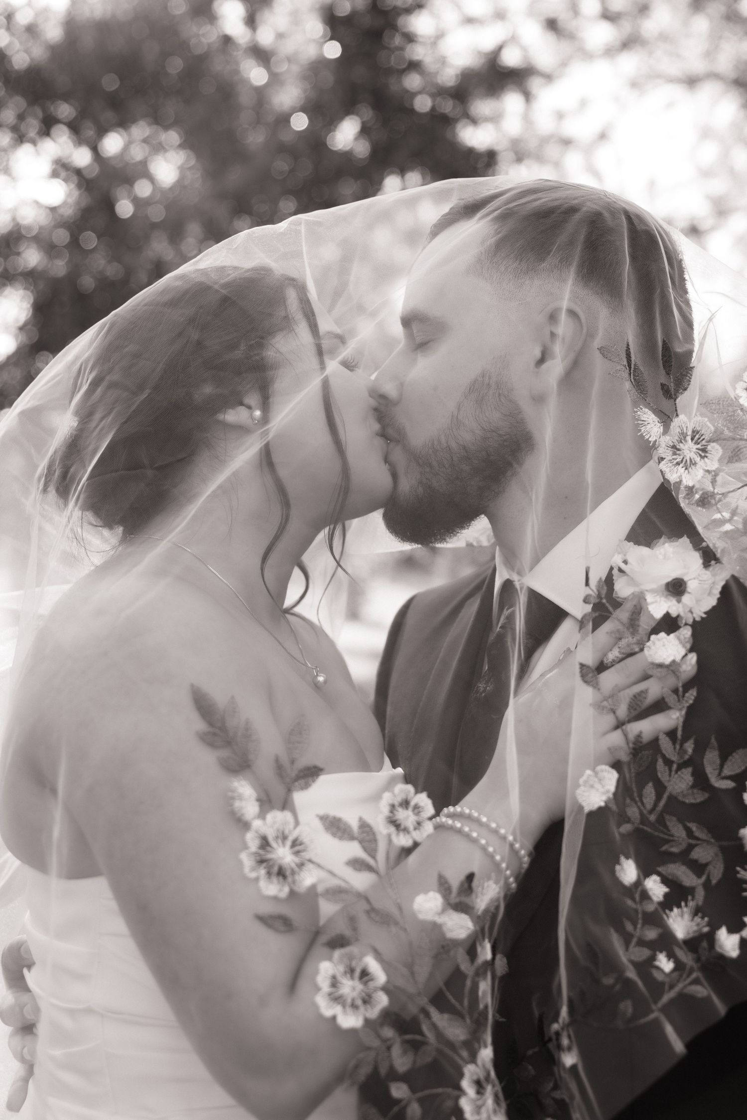 Black and white photo of a bride and groom kissing under a floral wedding veil outdoors.