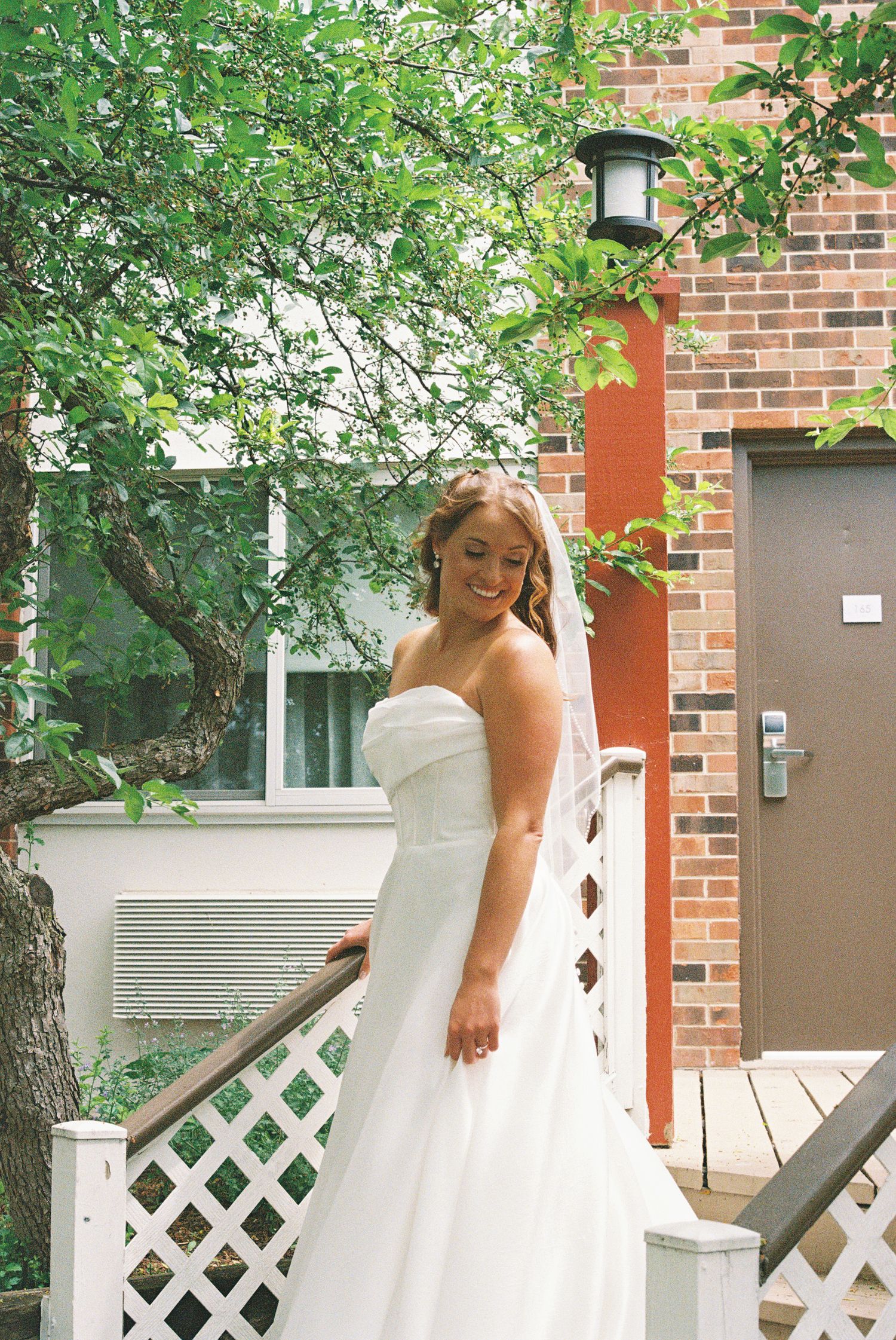 Smiling bride in strapless white wedding gown and veil standing on outdoor steps near brick building surrounded by green trees.