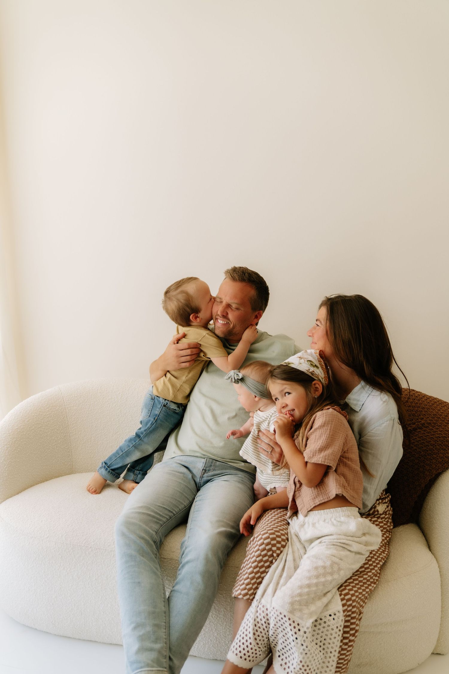 A happy family of four sits on a white sofa, with parents smiling as children cuddle close in a bright room.