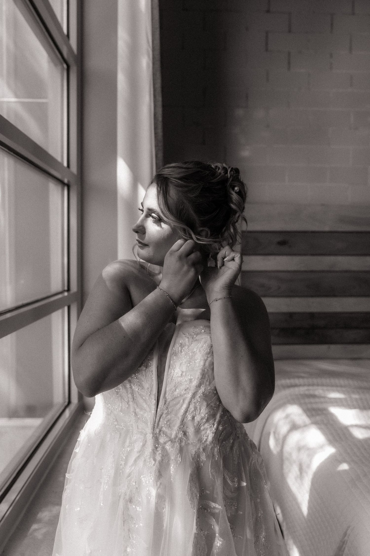 Black and white photo of a bride in a lace gown adjusting her earring by a sunlit window.