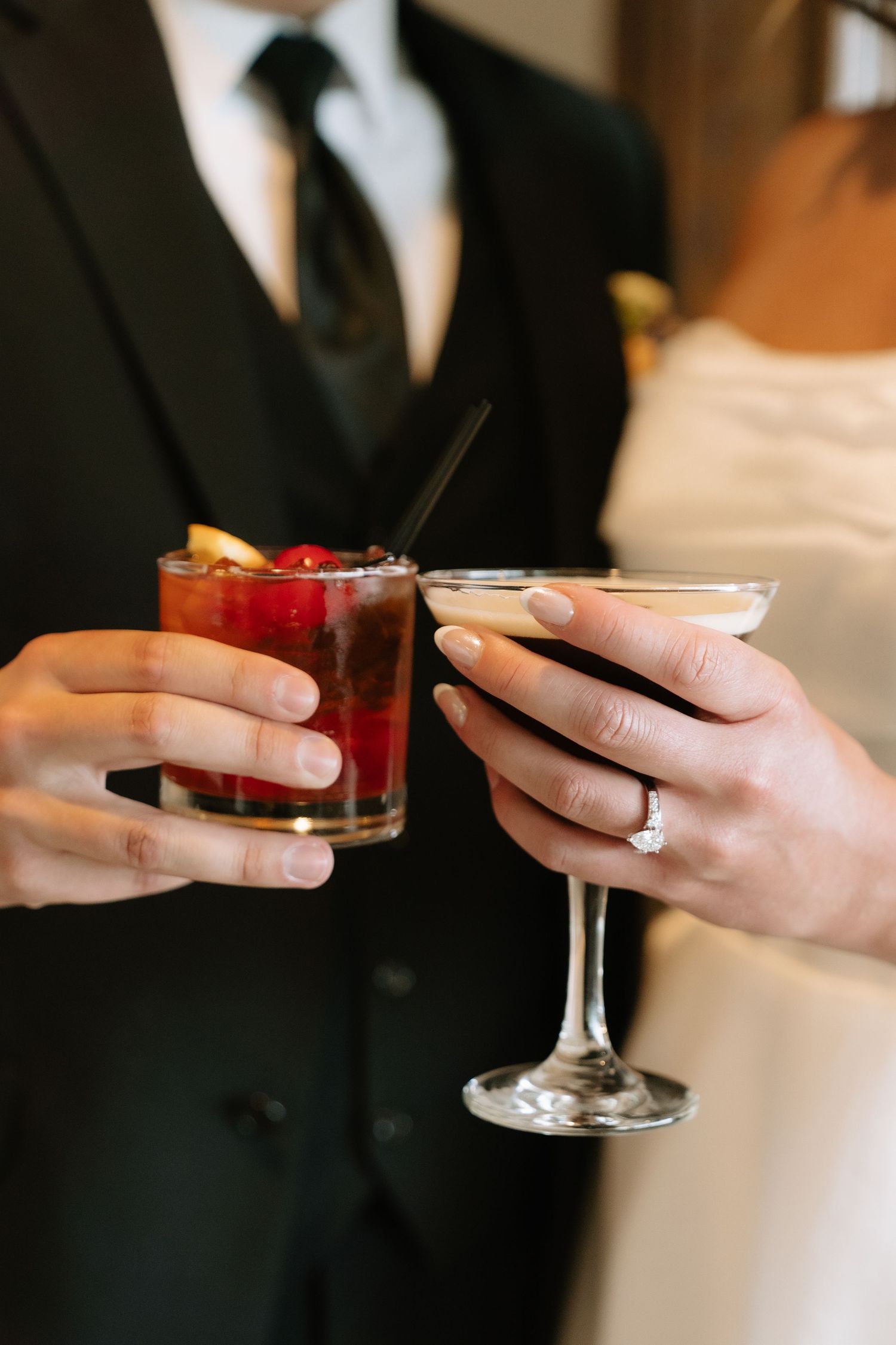 Bride and groom clinking cocktails, she wears an engagement ring, holding a martini and red berry drink.