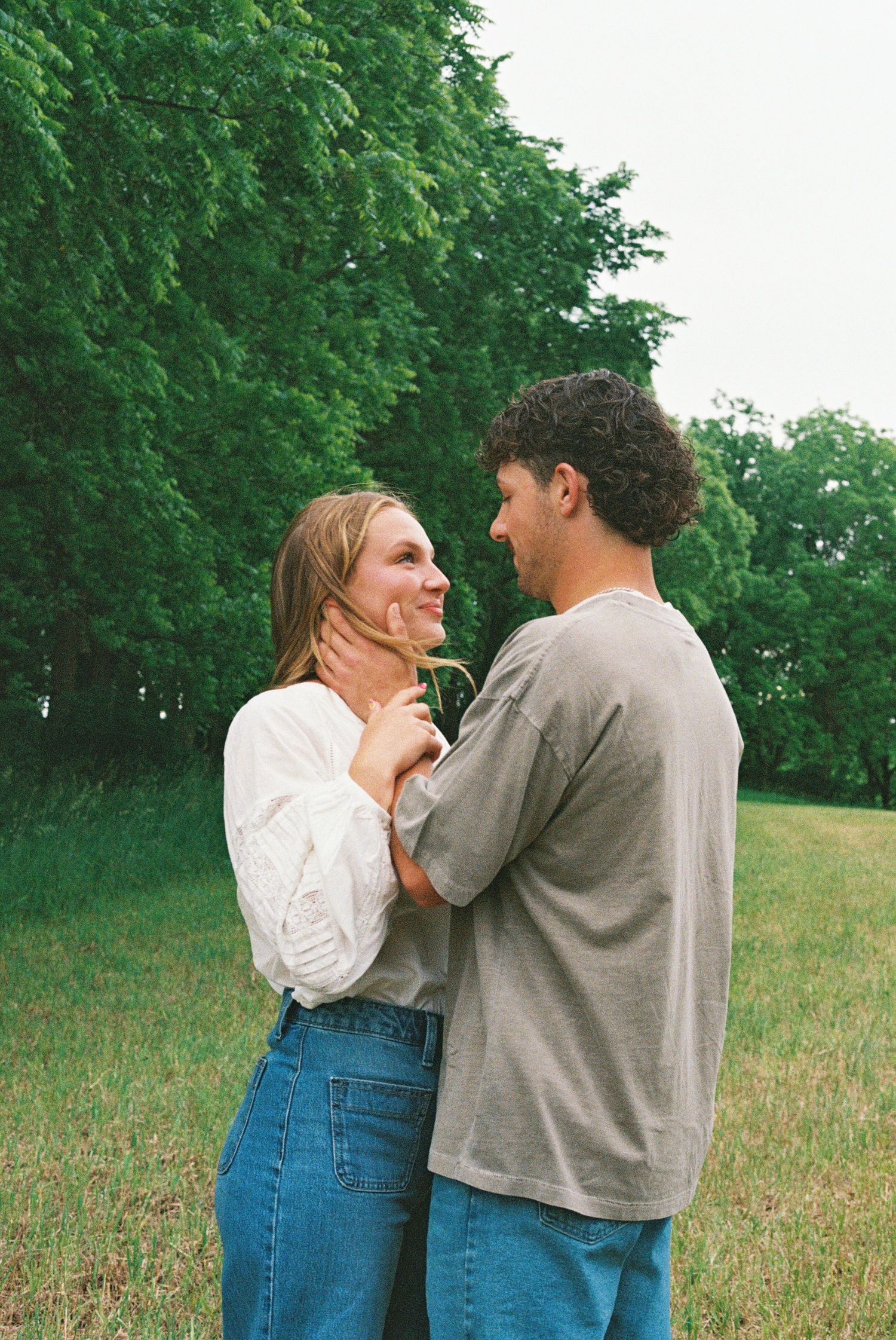 A couple shares a tender moment outdoors, facing each other on a green grass field with lush trees in the background.