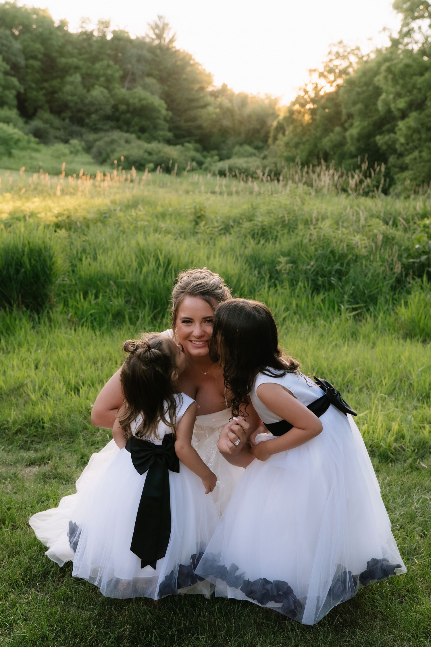 Bride in white gown crouching with two flower girls in white dresses with black bows in a lush green field.