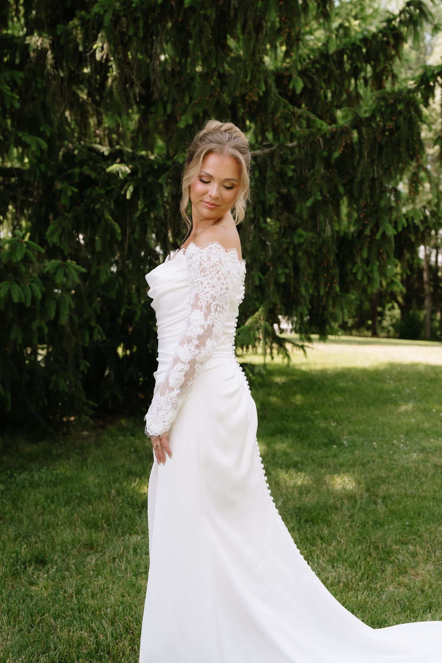 Bride in elegant white lace off-shoulder gown with long train poses outdoors near lush green evergreen trees.