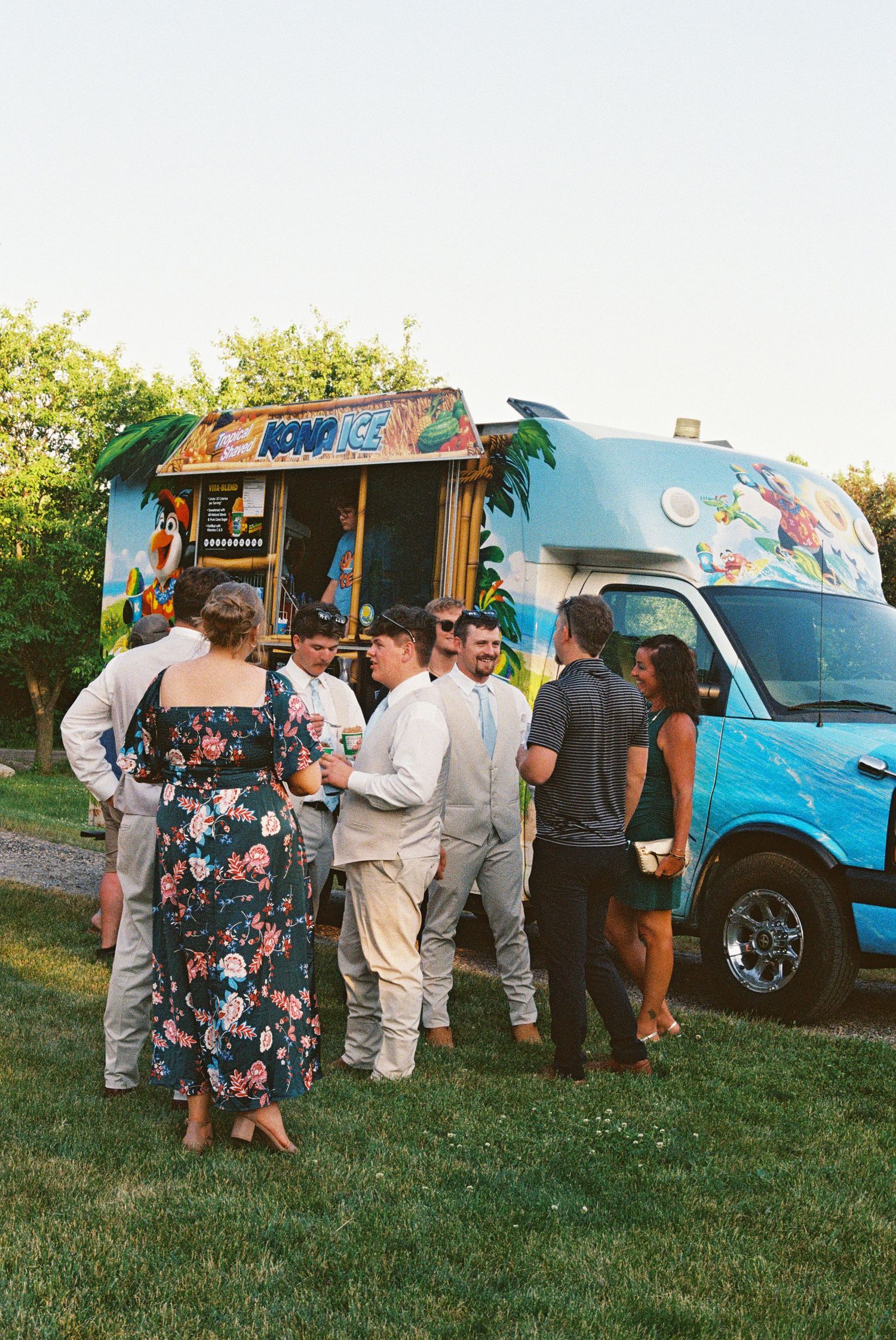 Guests gather around a colorful Kona Ice truck at an outdoor wedding reception on a sunny evening.
