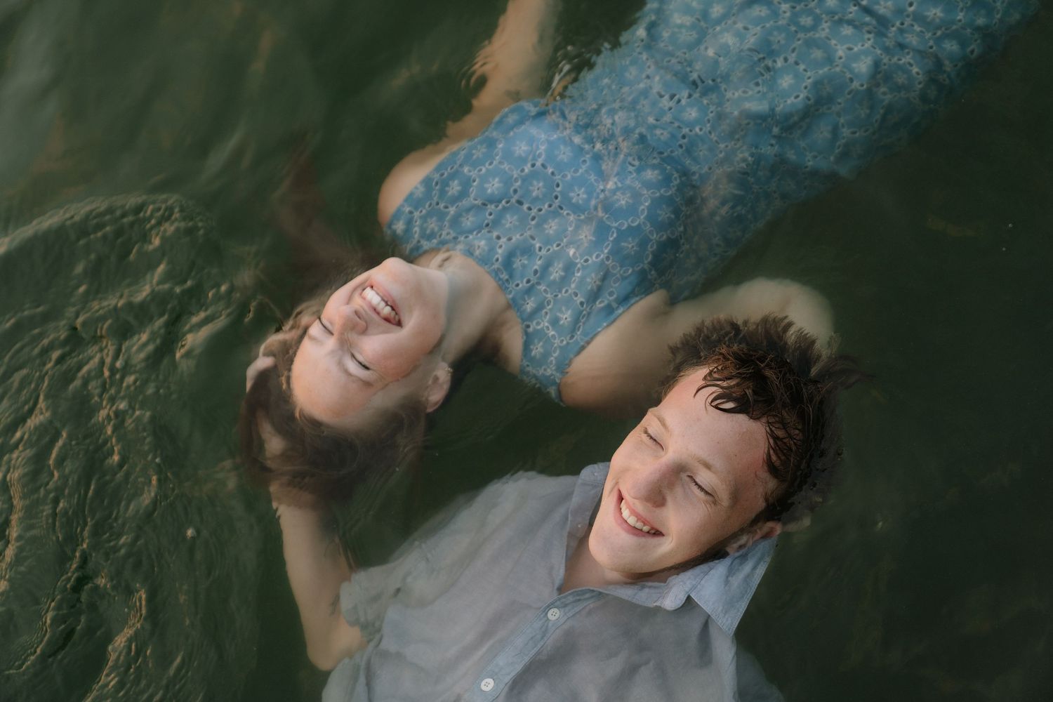 A smiling couple floating on their backs in calm water, wearing casual clothes, viewed from above.