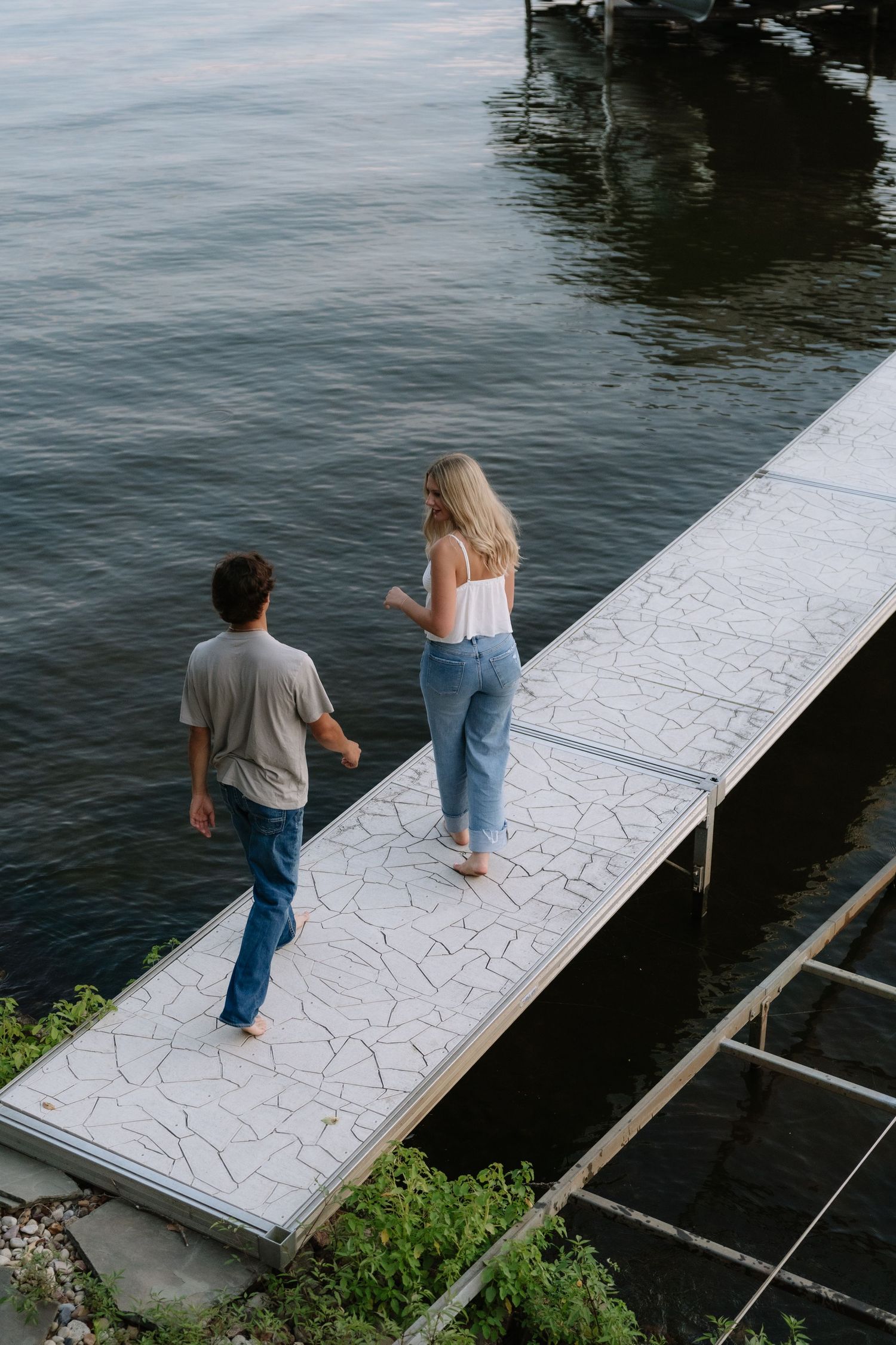 Couple walking barefoot on a white dock over calm water, wearing jeans and casual tops, viewed from above.