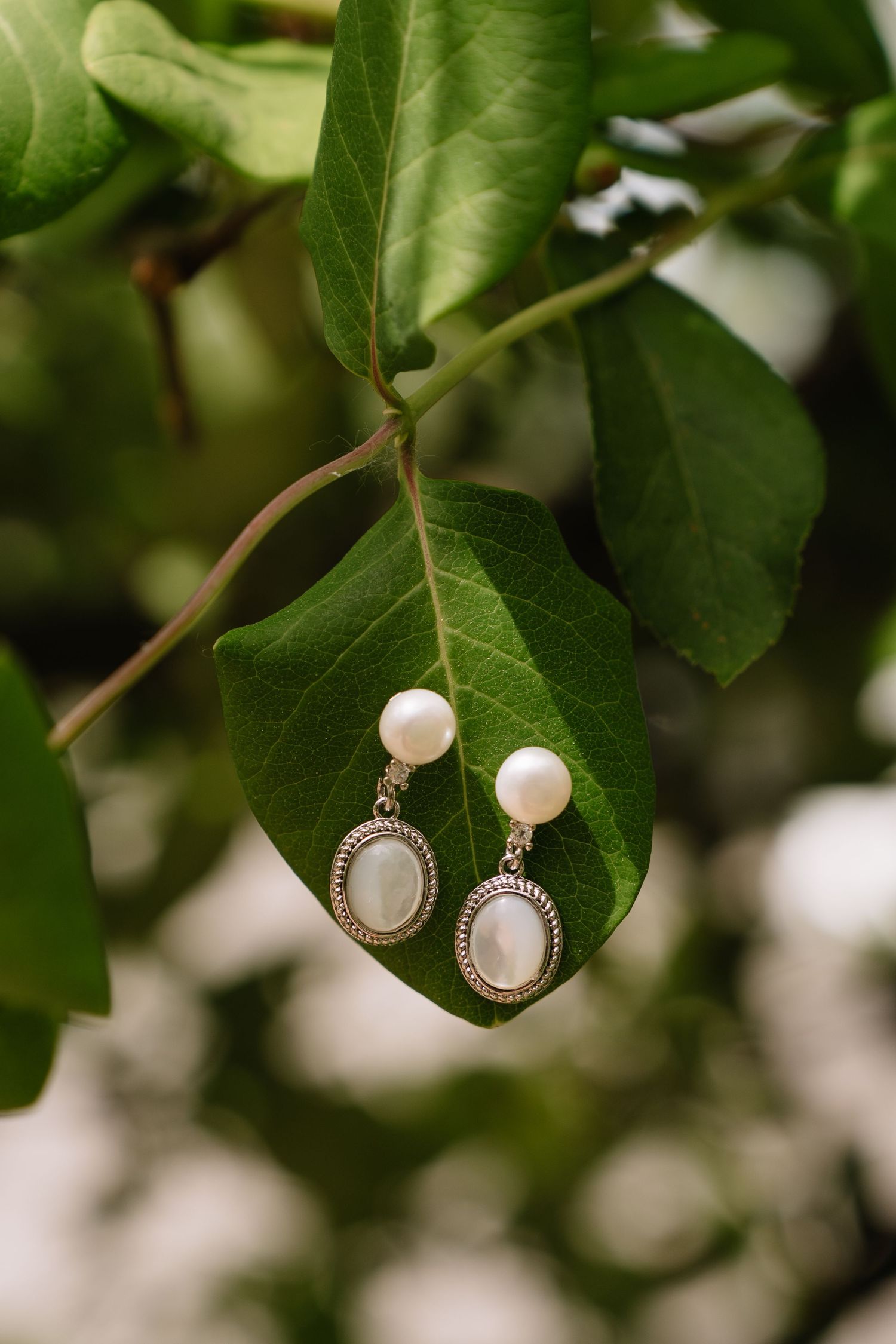 Elegant pearl drop earrings with mother-of-pearl oval pendants resting on a green leaf outdoors.