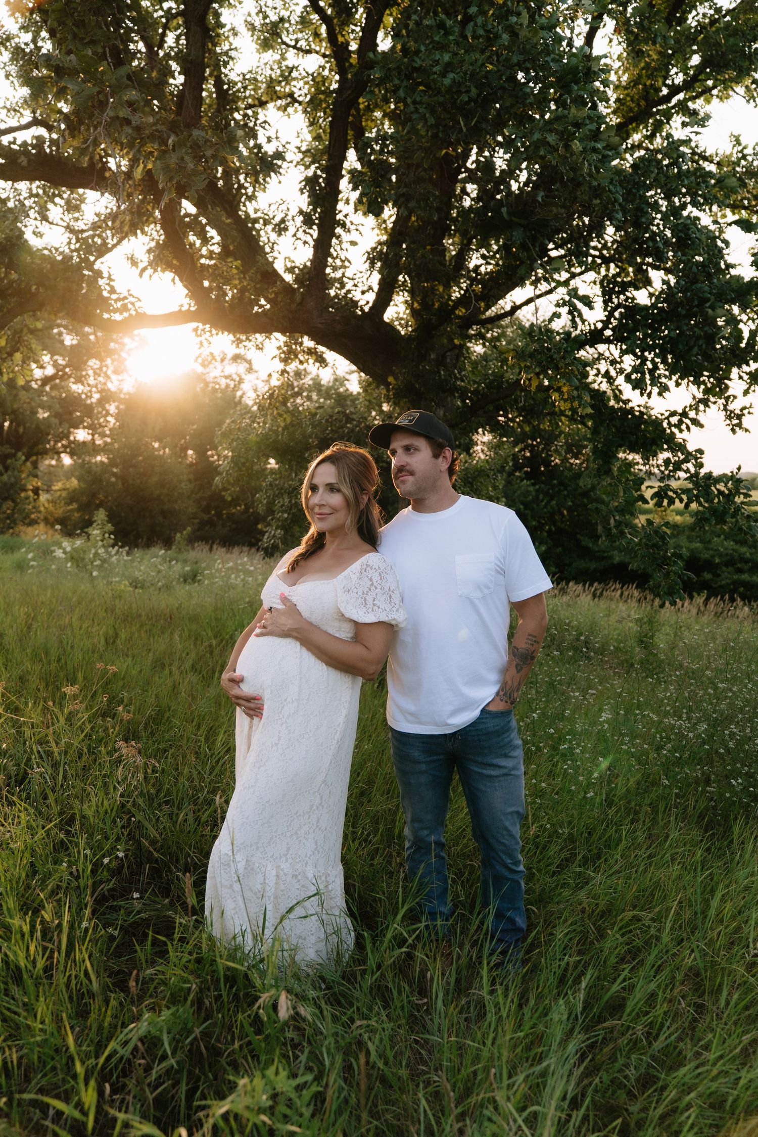 Pregnant woman in white lace dress poses with man in white tee and jeans at golden hour in lush green field.