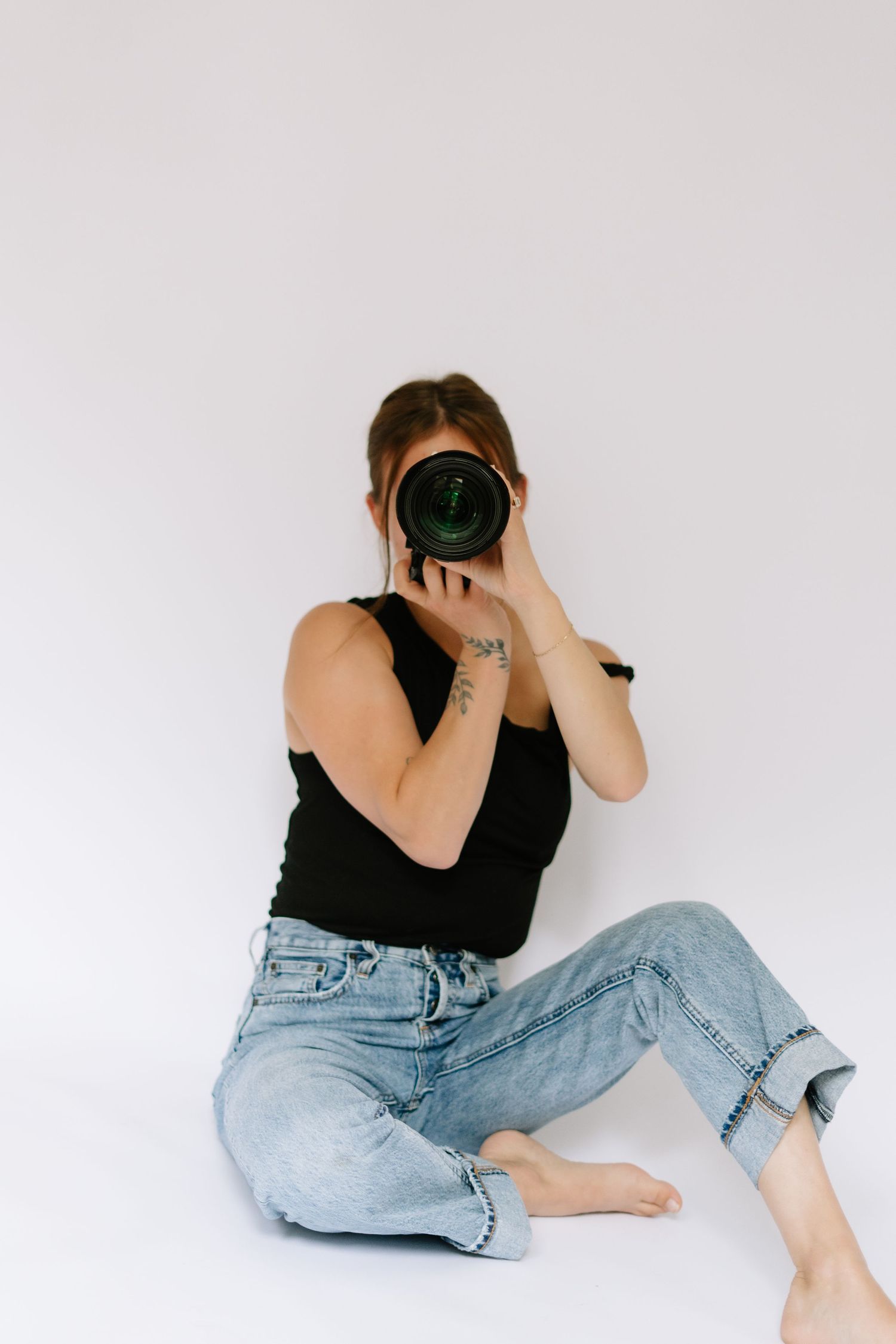 Female photographer sitting barefoot on floor aiming DSLR camera at viewer against white wall.