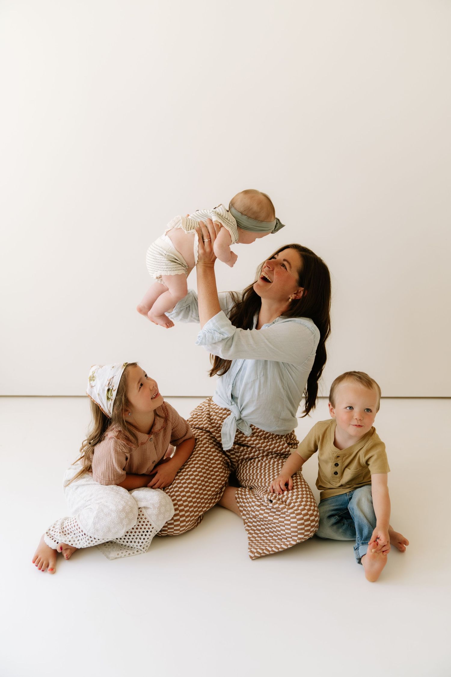 A joyful mother lifts her baby while two young children sit beside her on a white studio floor.