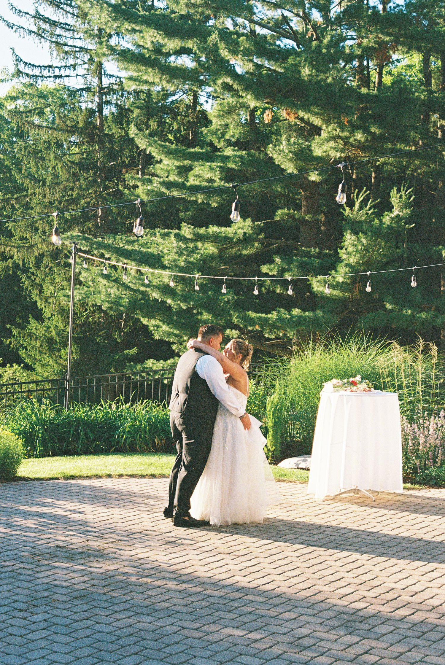 Bride and groom share first dance kiss on brick patio surrounded by lush green trees and string lights at outdoor wedding.