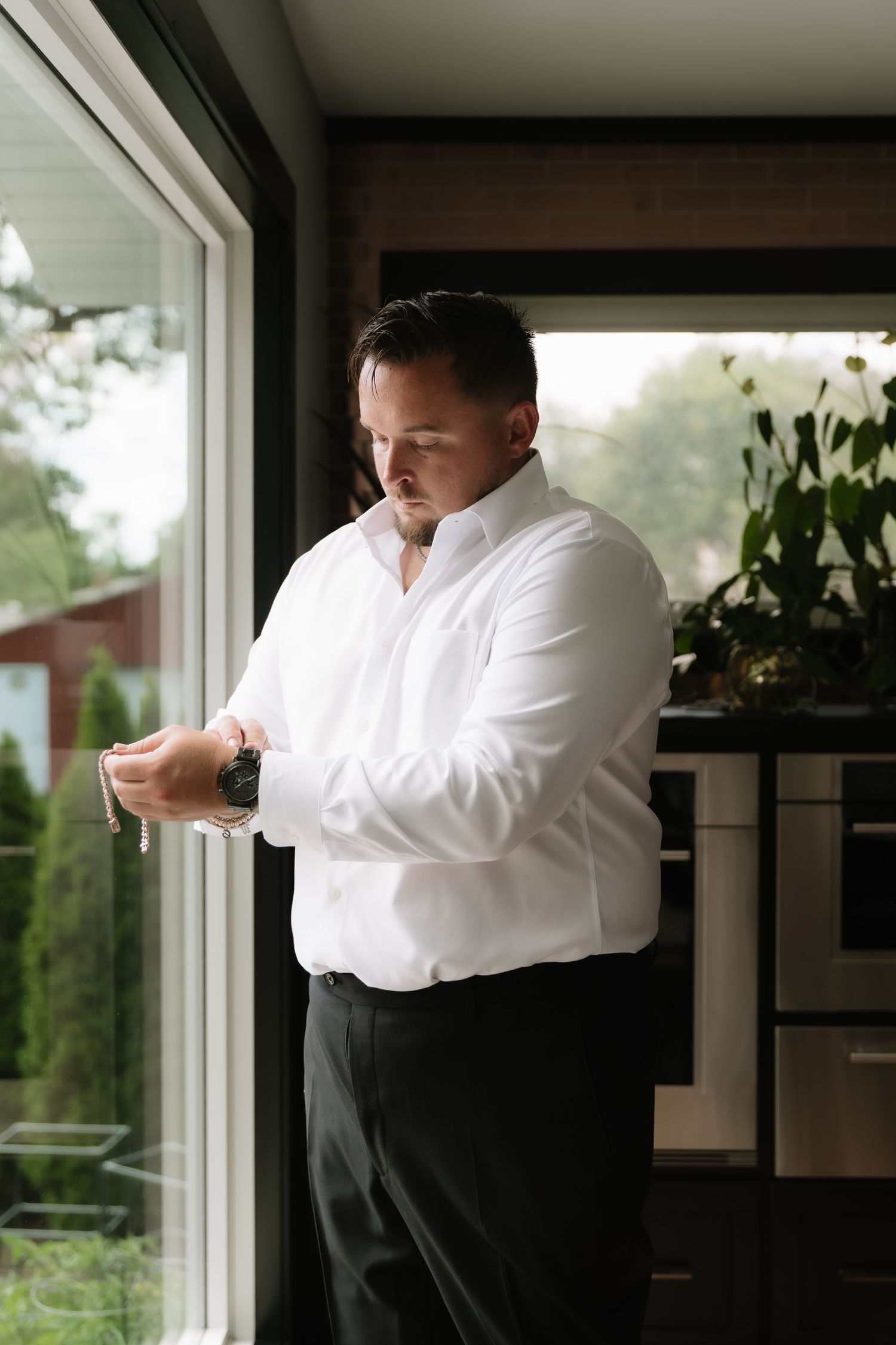 Man in white dress shirt adjusting cufflinks by window, wearing watch, dressed for formal occasion.