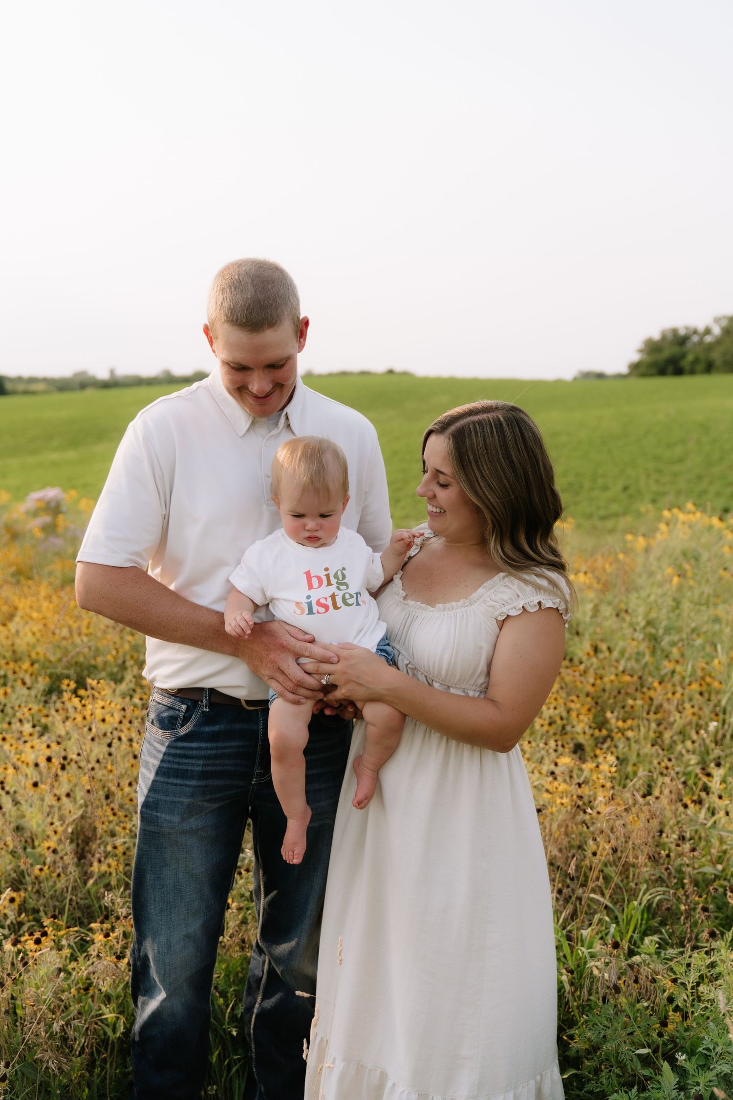 A young family poses in a wildflower field, baby wearing a big sister shirt, announcing a pregnancy.