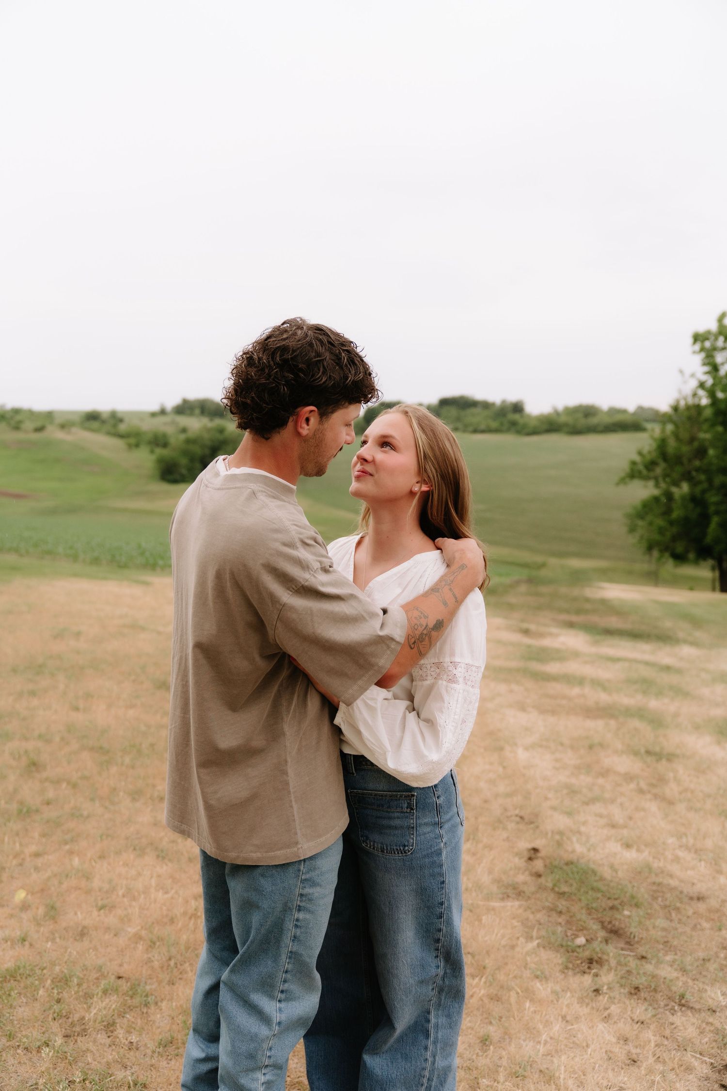 A couple embraces in a green field, the woman in a white blouse looking up at the man in a tan shirt and jeans.